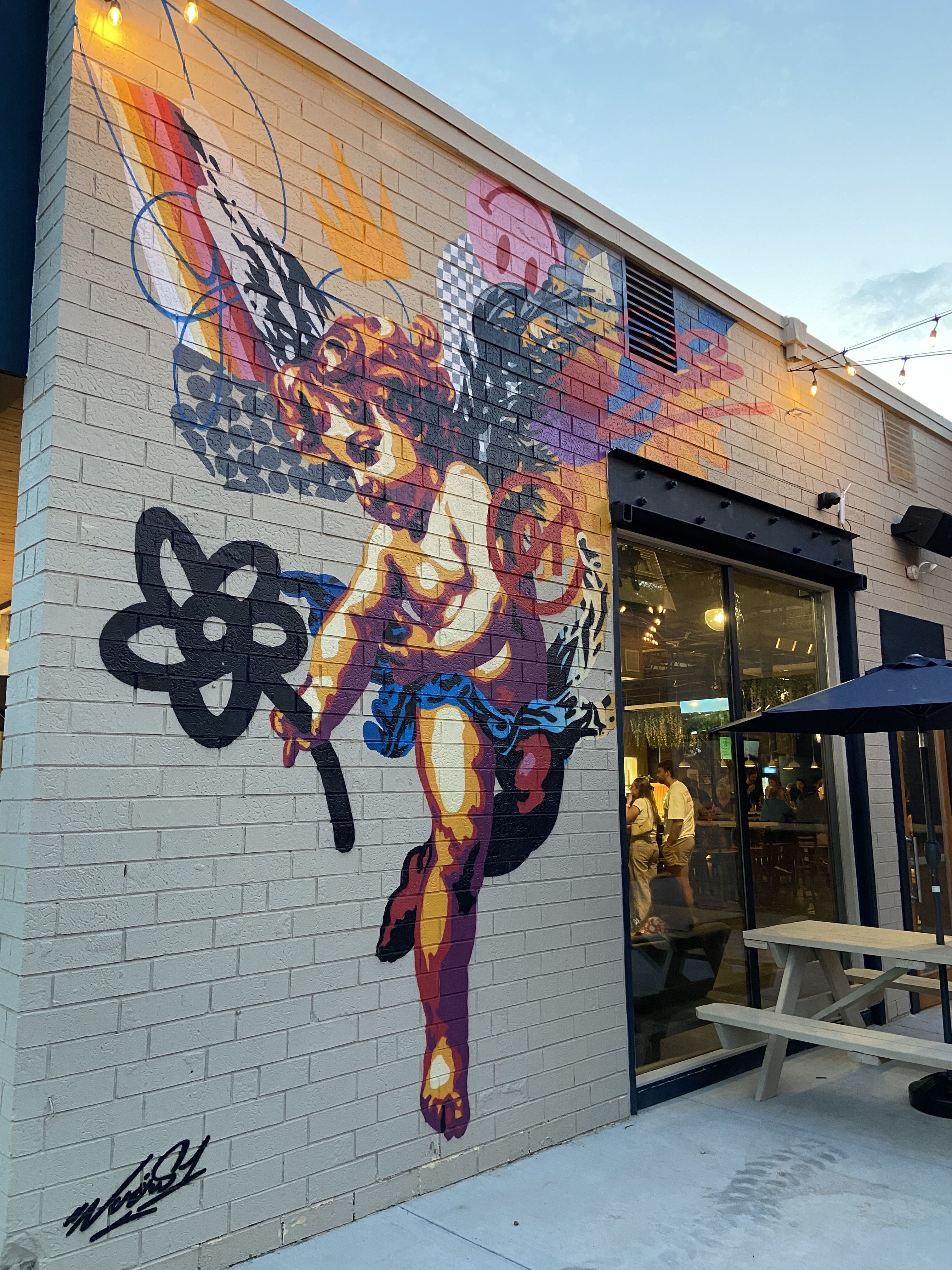 Colorful mural of a winged cherub holding a black flower on a brick wall next to a glass door with people inside and outdoor picnic tables under string lights at dusk.