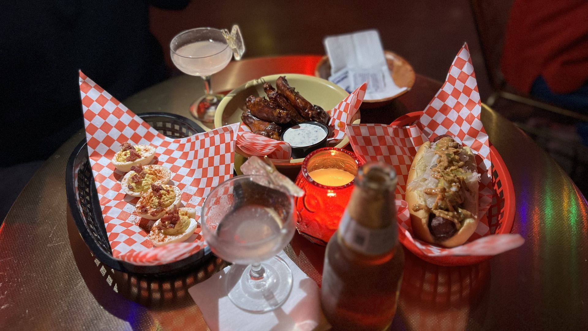 A table with deviled eggs, hot wings with dipping sauce, a hot dog with toppings, two cocktails—one with a butterfly garnish—and a beer bottle in a dimly lit setting.