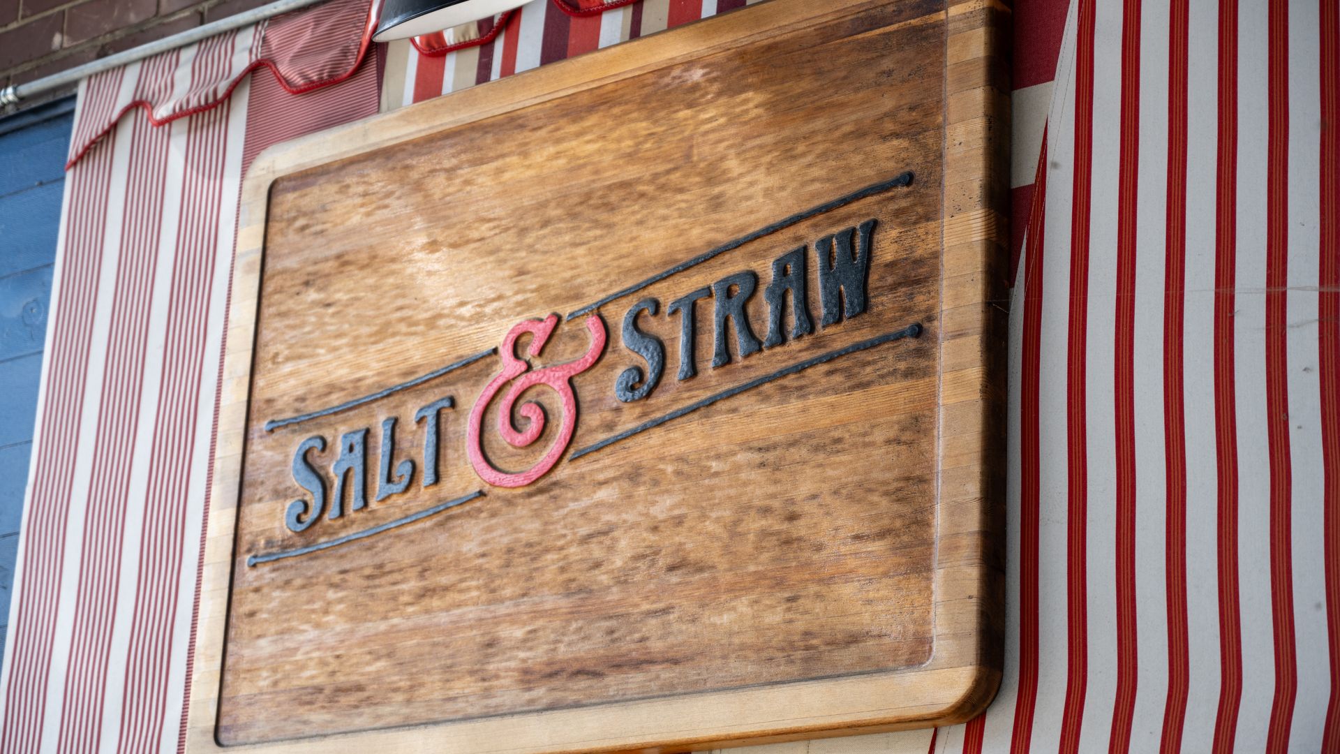 Weathered wooden sign reading "Salt & Straw" with a pink ampersand, set against a red-and-white striped storefront backdrop.