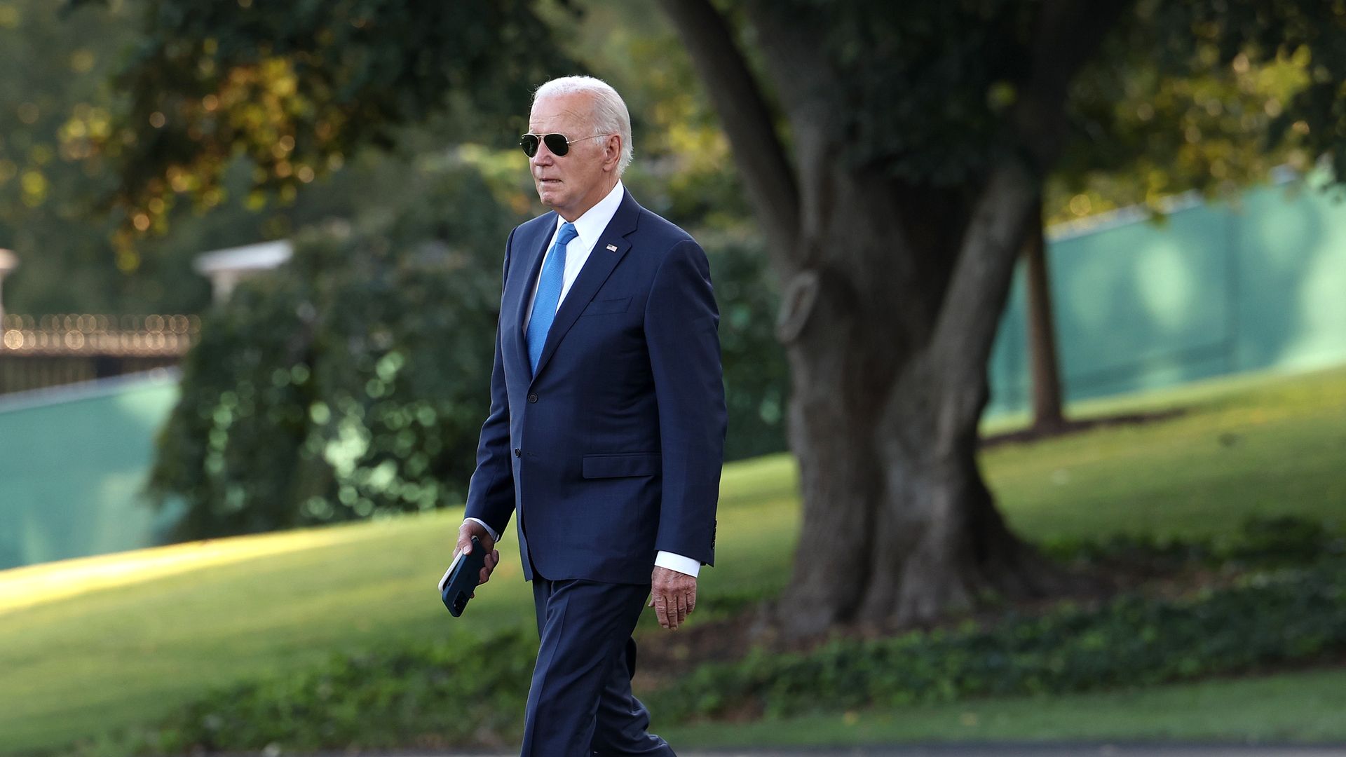 President Biden walks across the South Lawn of the White House, wearing a blue suit and sunglasses.