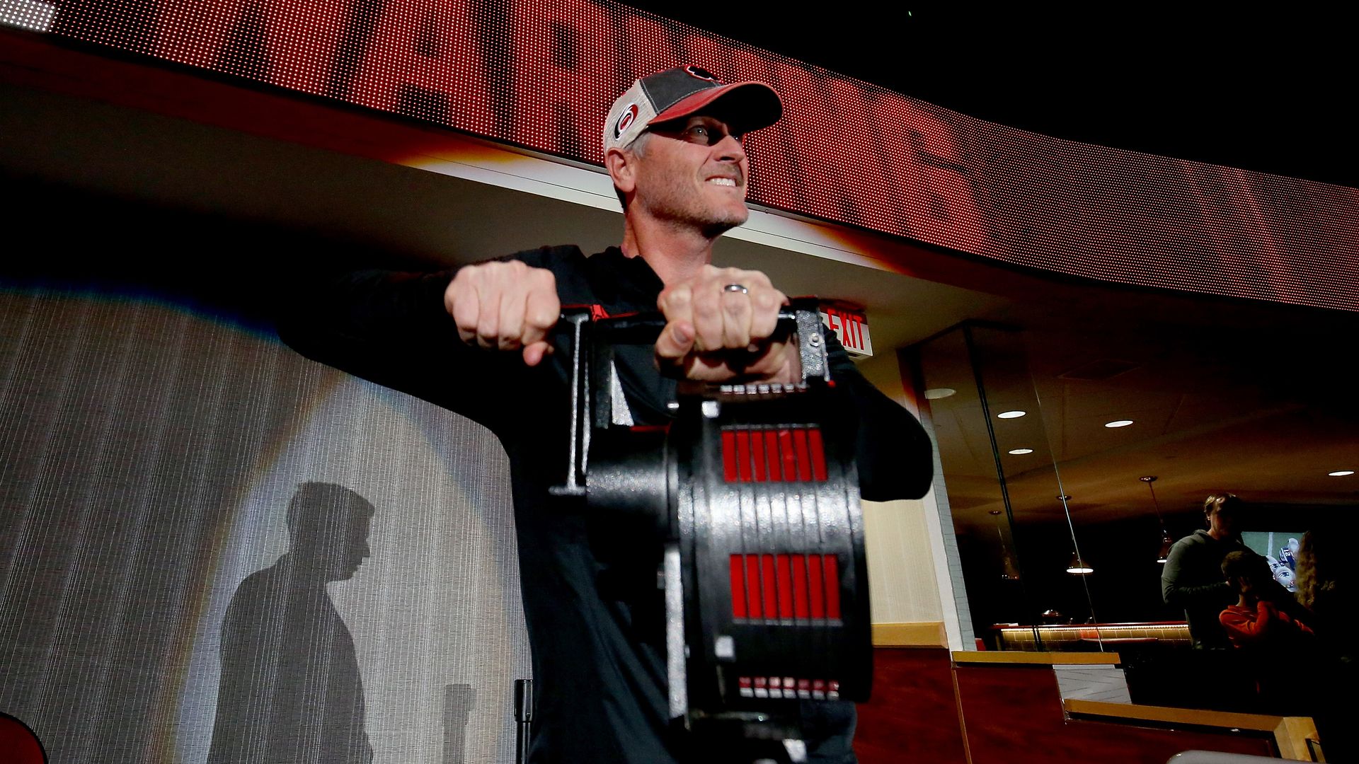 Man in black shirt and cap gripping a large black and red mechanical device indoors with shadow on wall, digital red and black sign above, and people in background.
