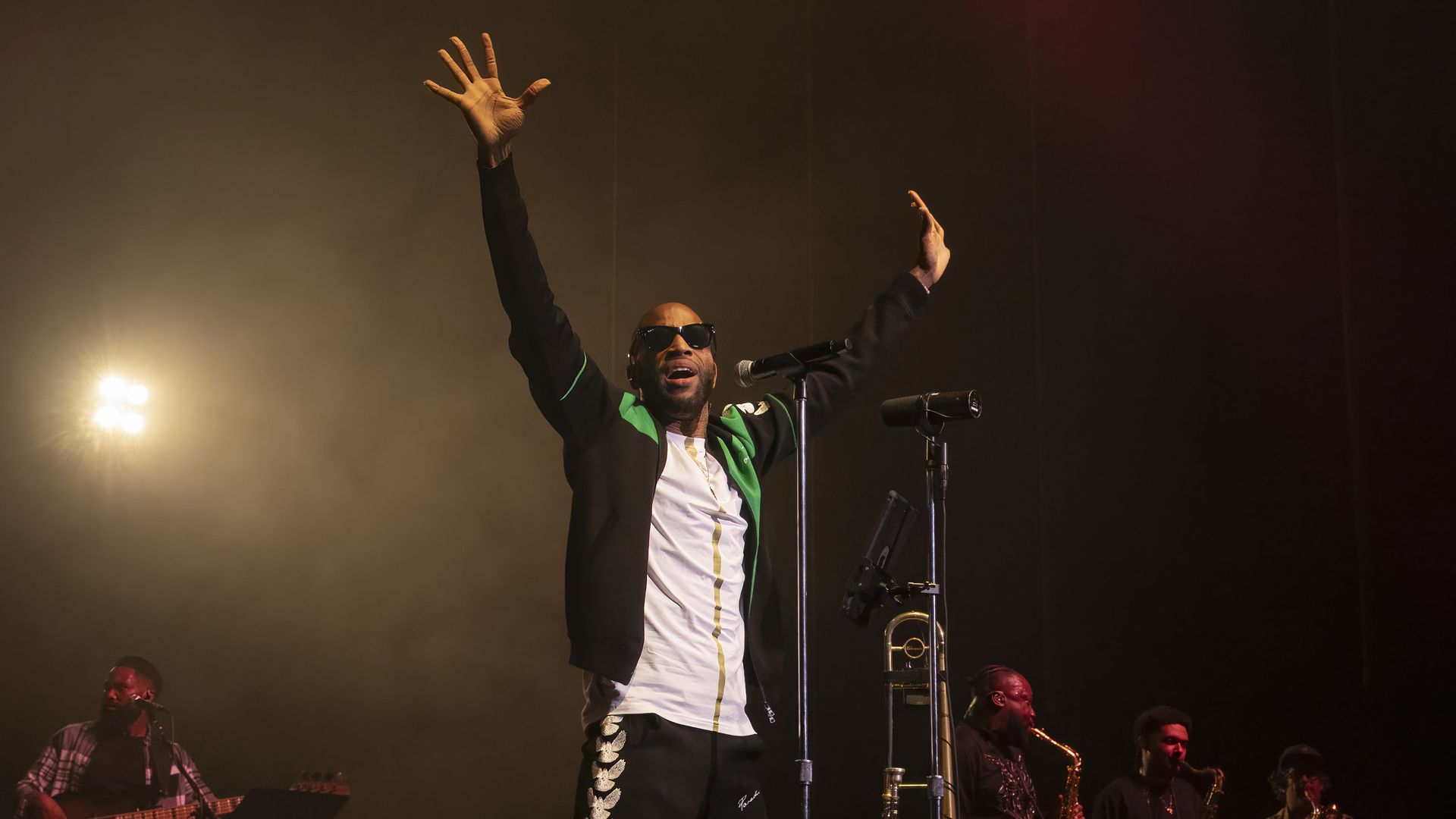 Trombone Shorty stands on stage next to a microphone with both arms stretched out above him.