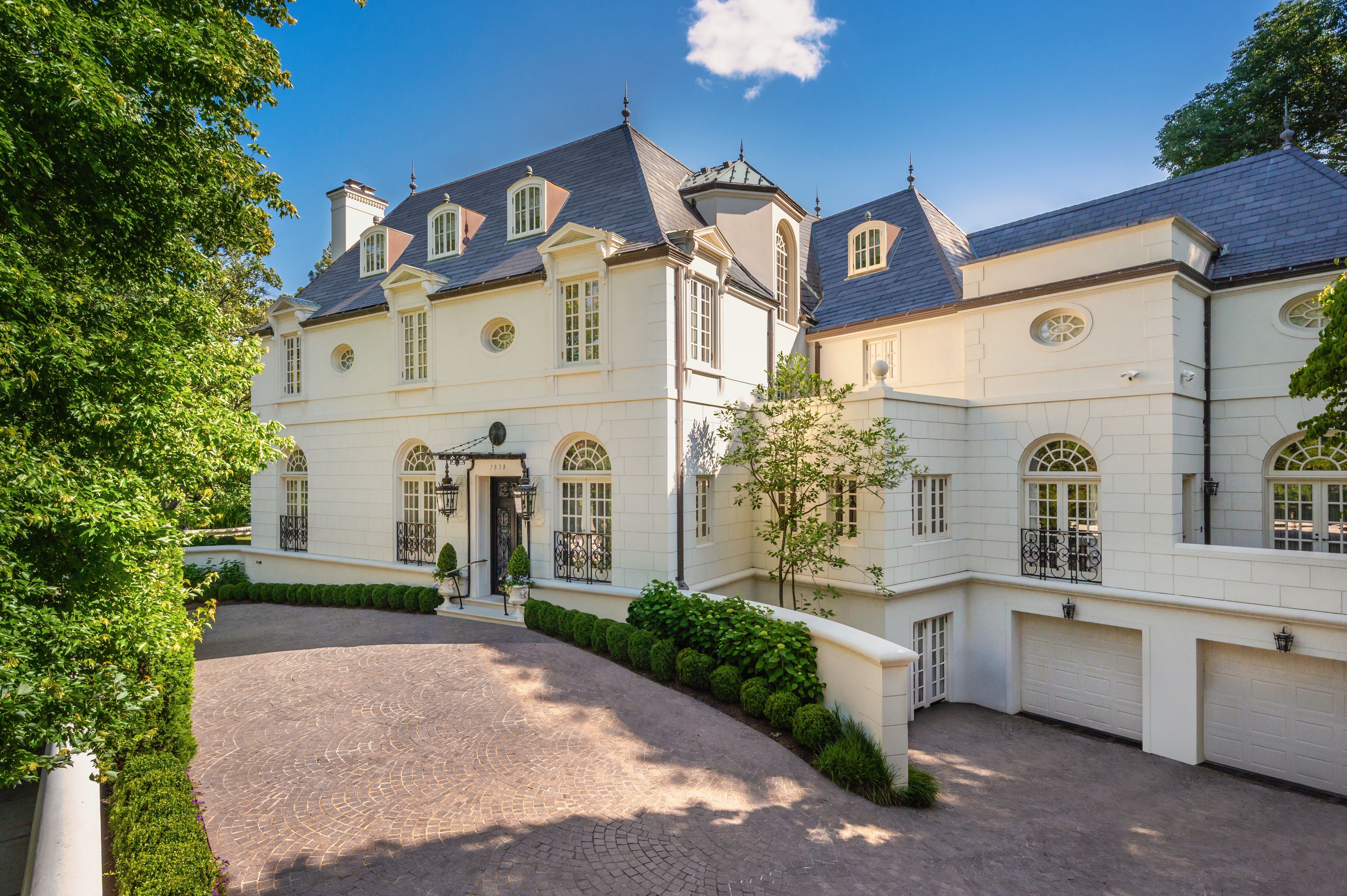 Large elegant white mansion with slate roof, arched windows, and black wrought-iron details, surrounded by green trees and bushes, under a blue sky with a few clouds.