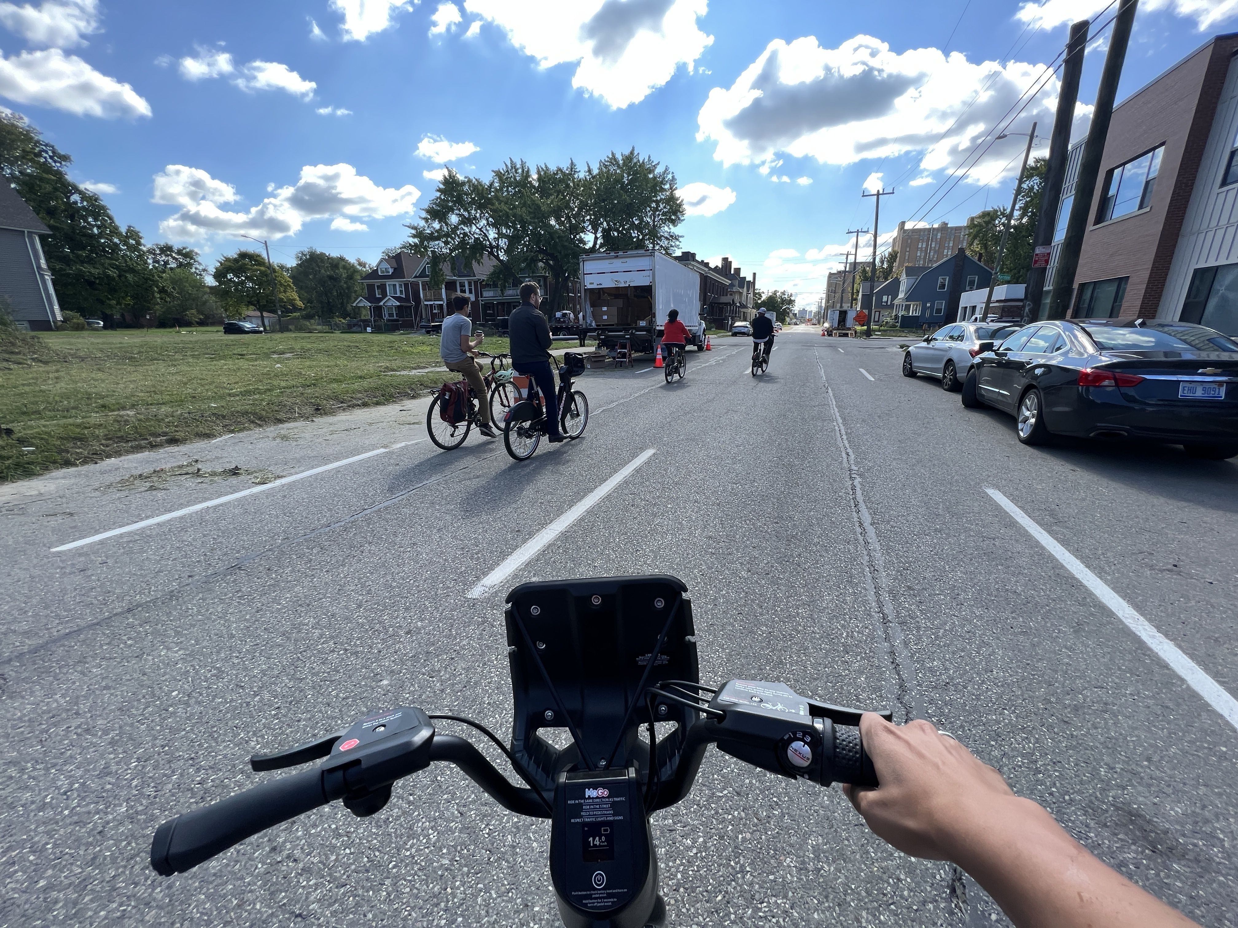 A picture of a MoGo e-bike shown from the perspective of the person riding it. They're rolling down a street with other bikes in front, sun shining, blue sky.