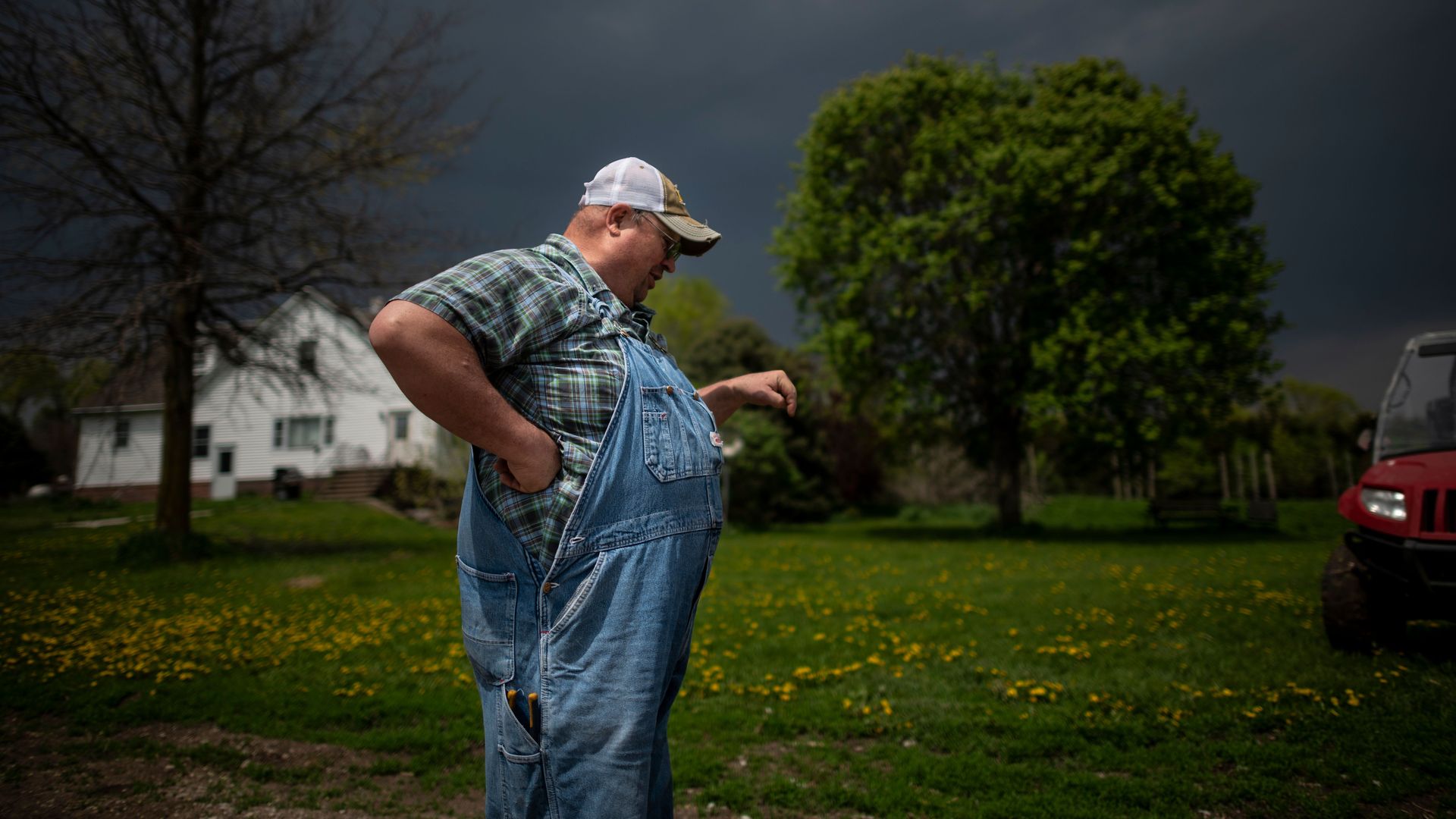 In this image, a farmer in overalls stands in a field with a white house in the background.