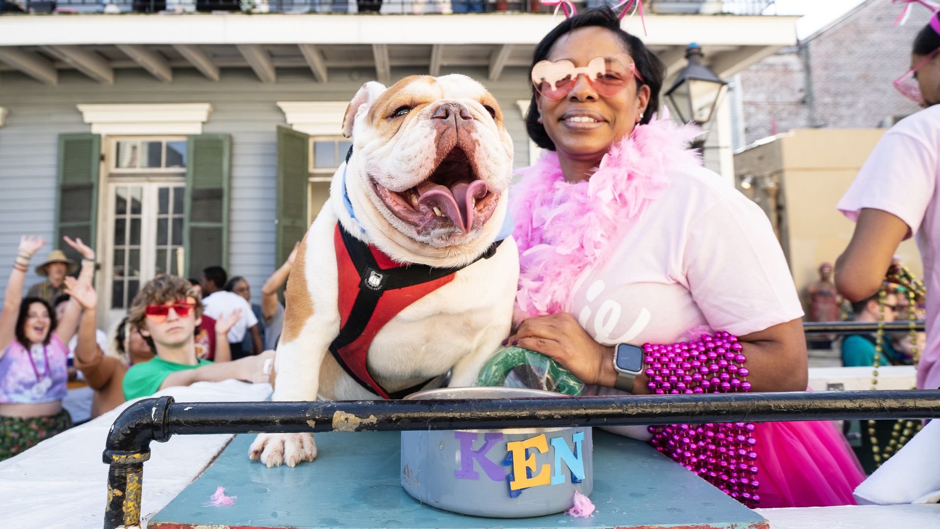 A bulldog sits on a platform next to a dog bowl that says KEN as a woman leans in to smile for the camera.