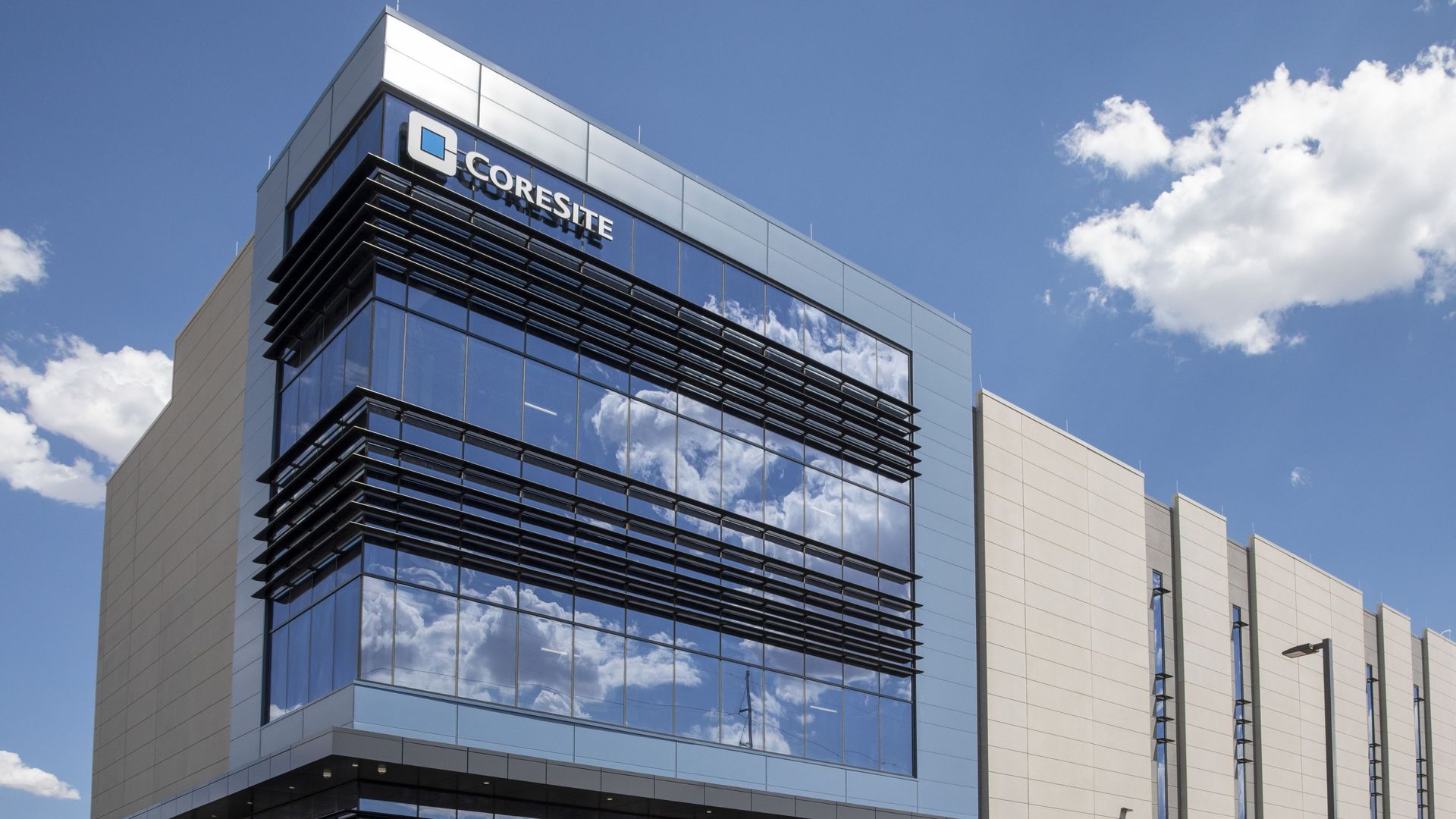 Modern glass office building with blue-tinted windows, black sunshades, and a CoreSite sign; blue sky with white clouds, a fenced perimeter, and a concrete plaque in the foreground.