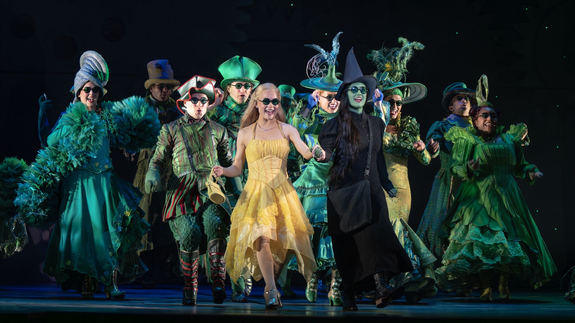 Group of performers on a dark stage wearing vibrant costumes and hats, predominantly green and gold, smiling and posing together under stage lighting.
