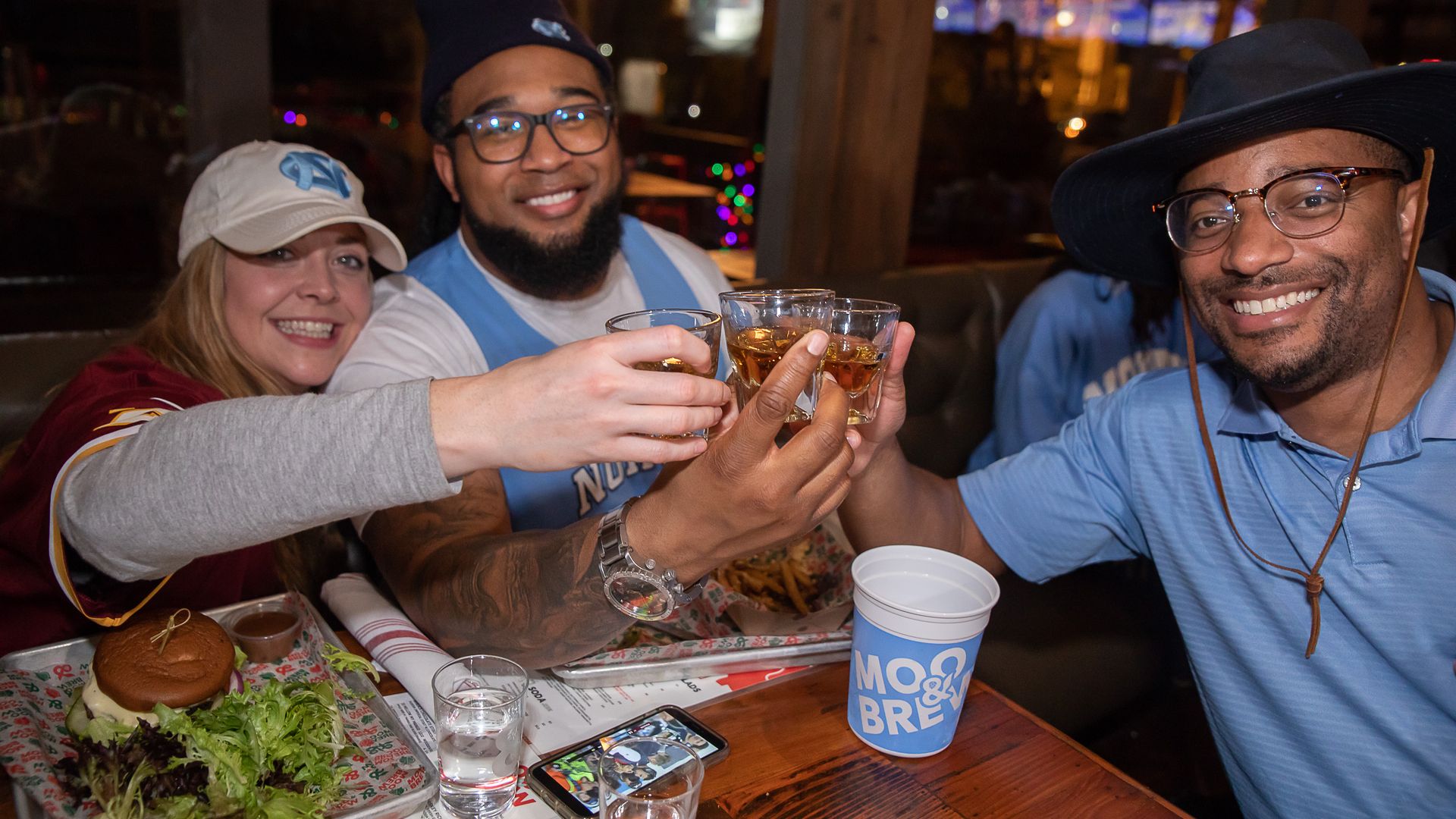 Three friends smiling and toasting with drinks while seated at a table with food, wearing casual clothes and hats in a dimly lit restaurant or bar.