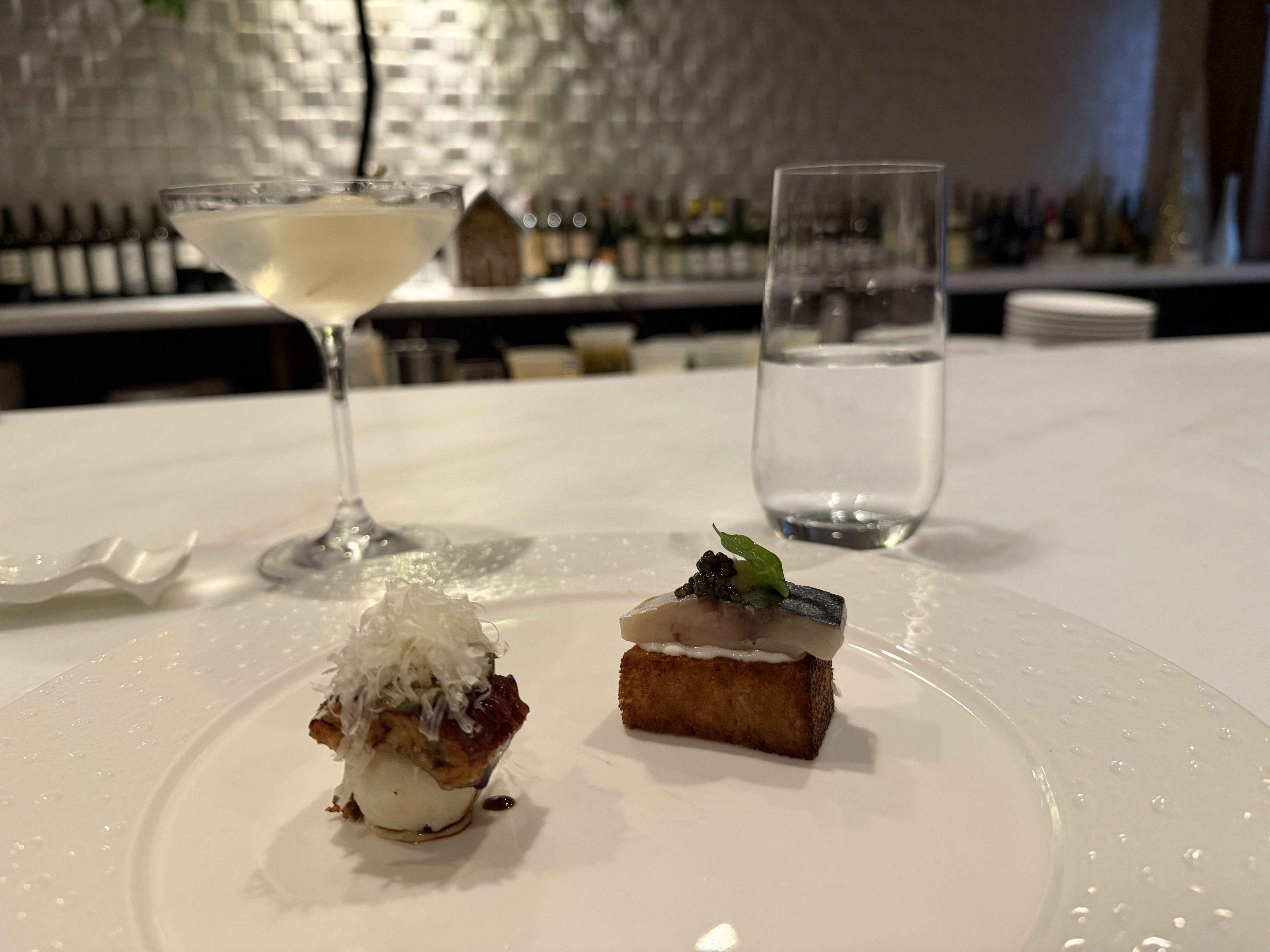 A white plate with two gourmet bite-sized appetizers on a white countertop. To the left, a cocktail glass with a pale yellow drink and a dark straw. To the right, a clear glass of water.
