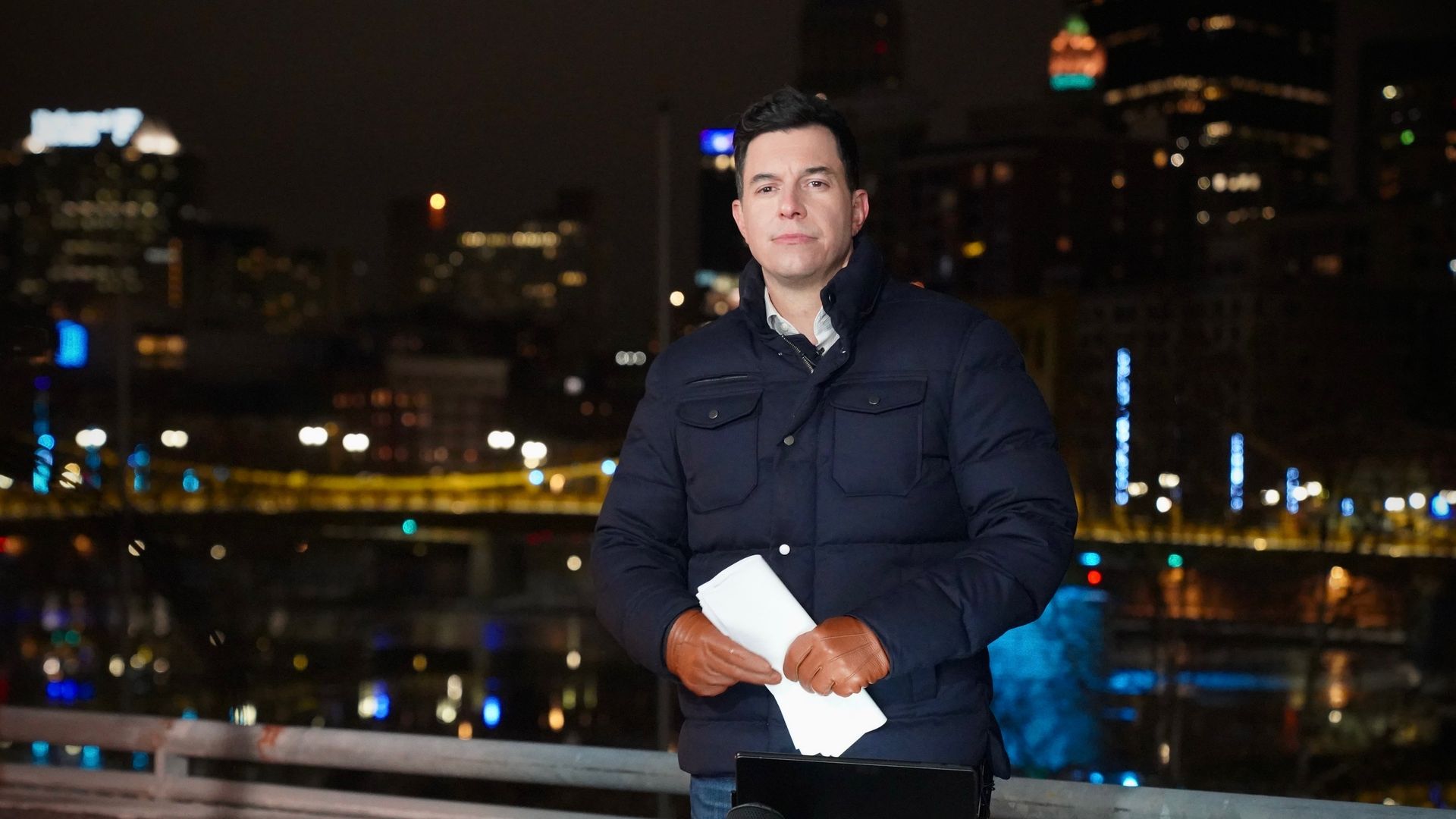 A man in a dark blue jacket and brown gloves stands holding white papers at night with a city skyline and blurred lights in the background, next to a laptop.