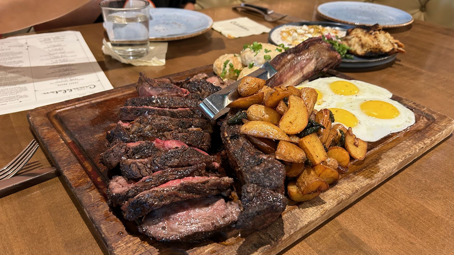 Wooden serving board with sliced medium-rare steak, crispy roasted potatoes, and two sunny-side-up eggs; plates and a glass on a restaurant table in the background.