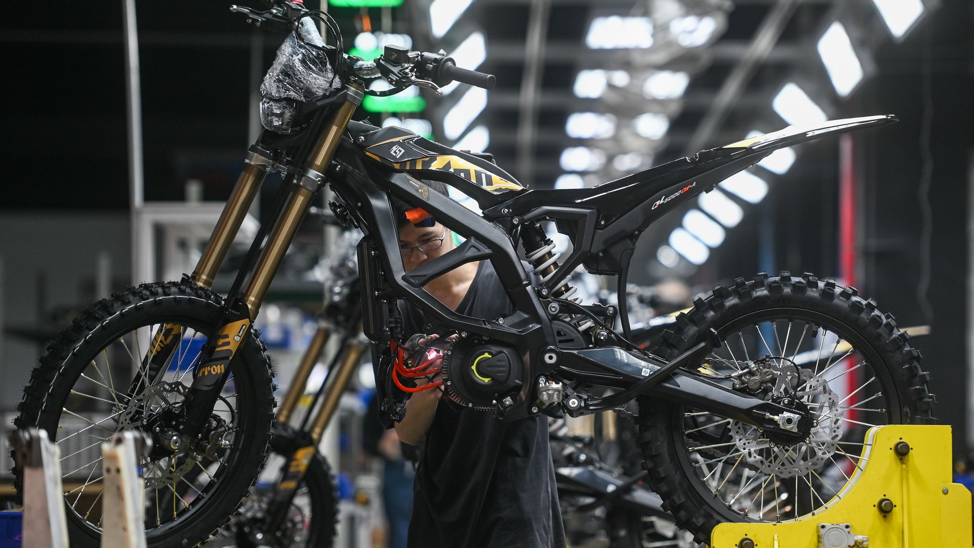 Mechanic in dark clothing works on a black and yellow dirt bike mounted on a yellow stand in a workshop. Gold front forks, knobby tires, and bright overhead lights show in the background.