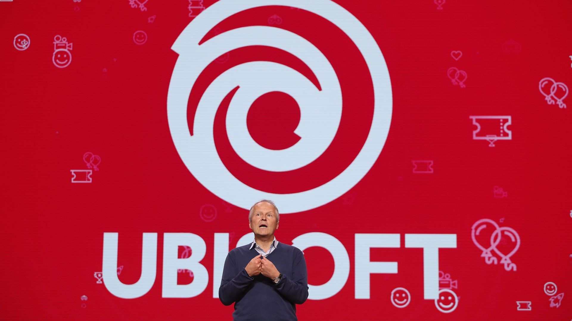 Photo of a man in pants, sweater, and a collard shirt standing in front of a big version of the Ubisoft logo