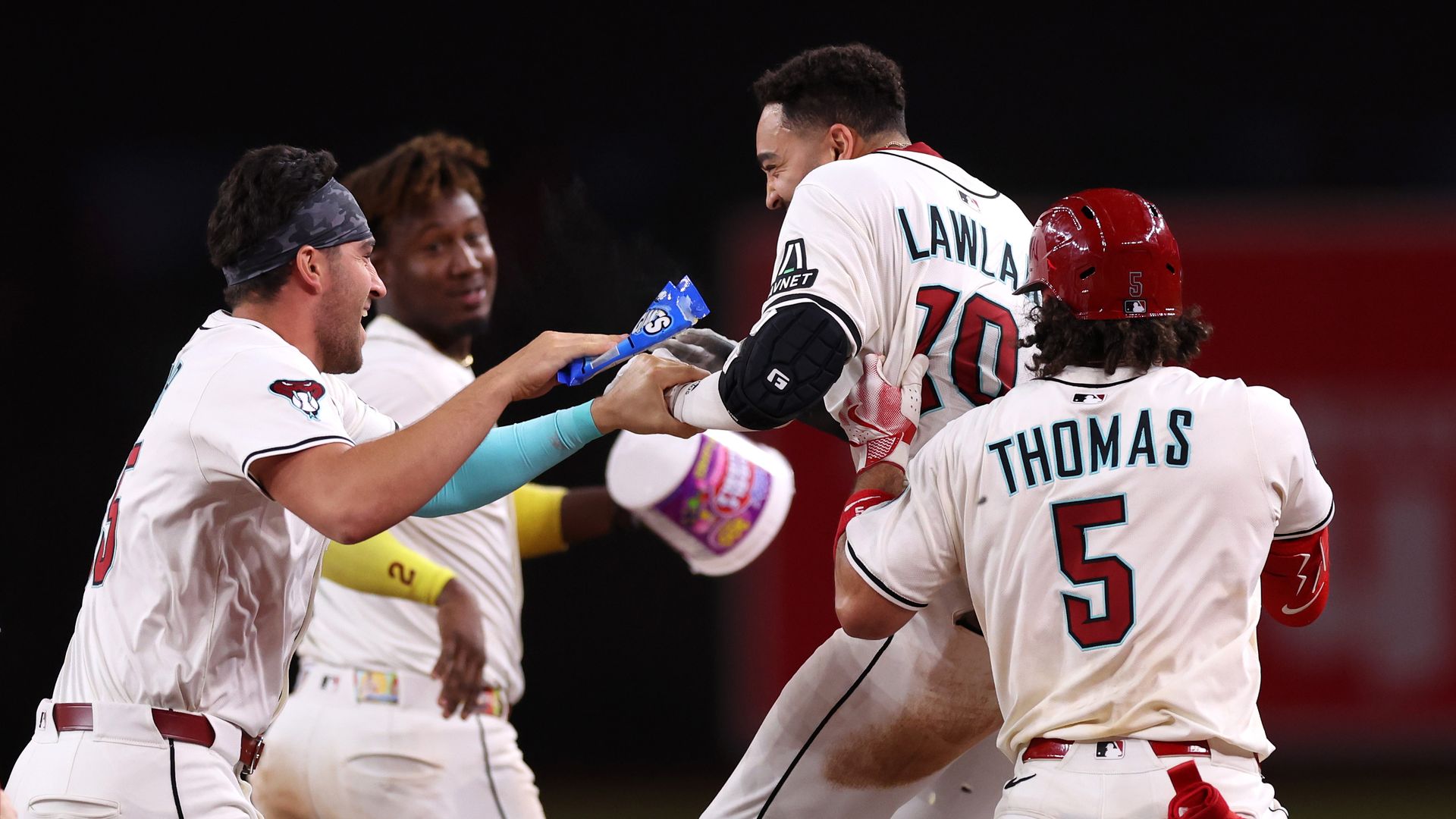 Four baseball players in white uniforms, with one wearing a red helmet, celebrate together. 