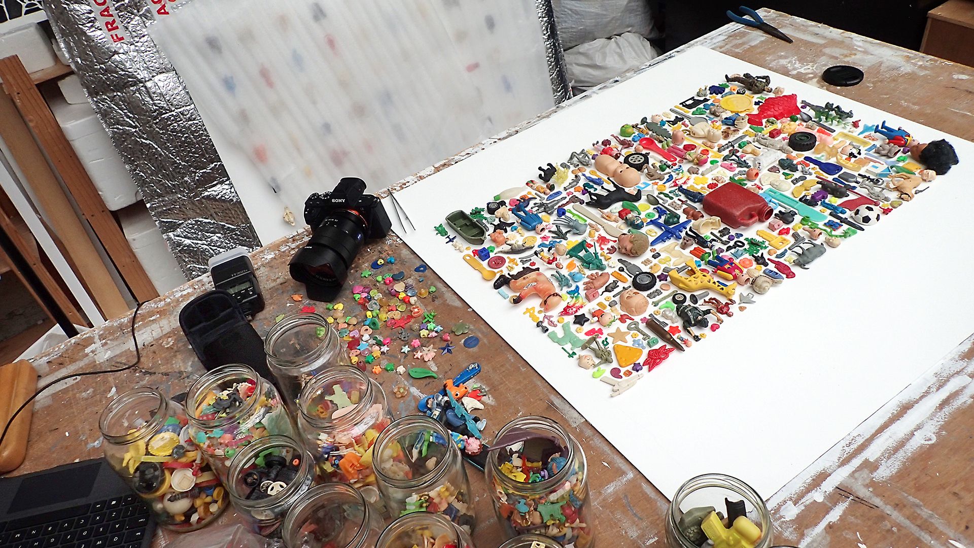 Images of artist's workspace with many glass jars holding assorted small plastic items like toys and beads. Next to it, a canvas with plastic objects arranged in a colorful montage
