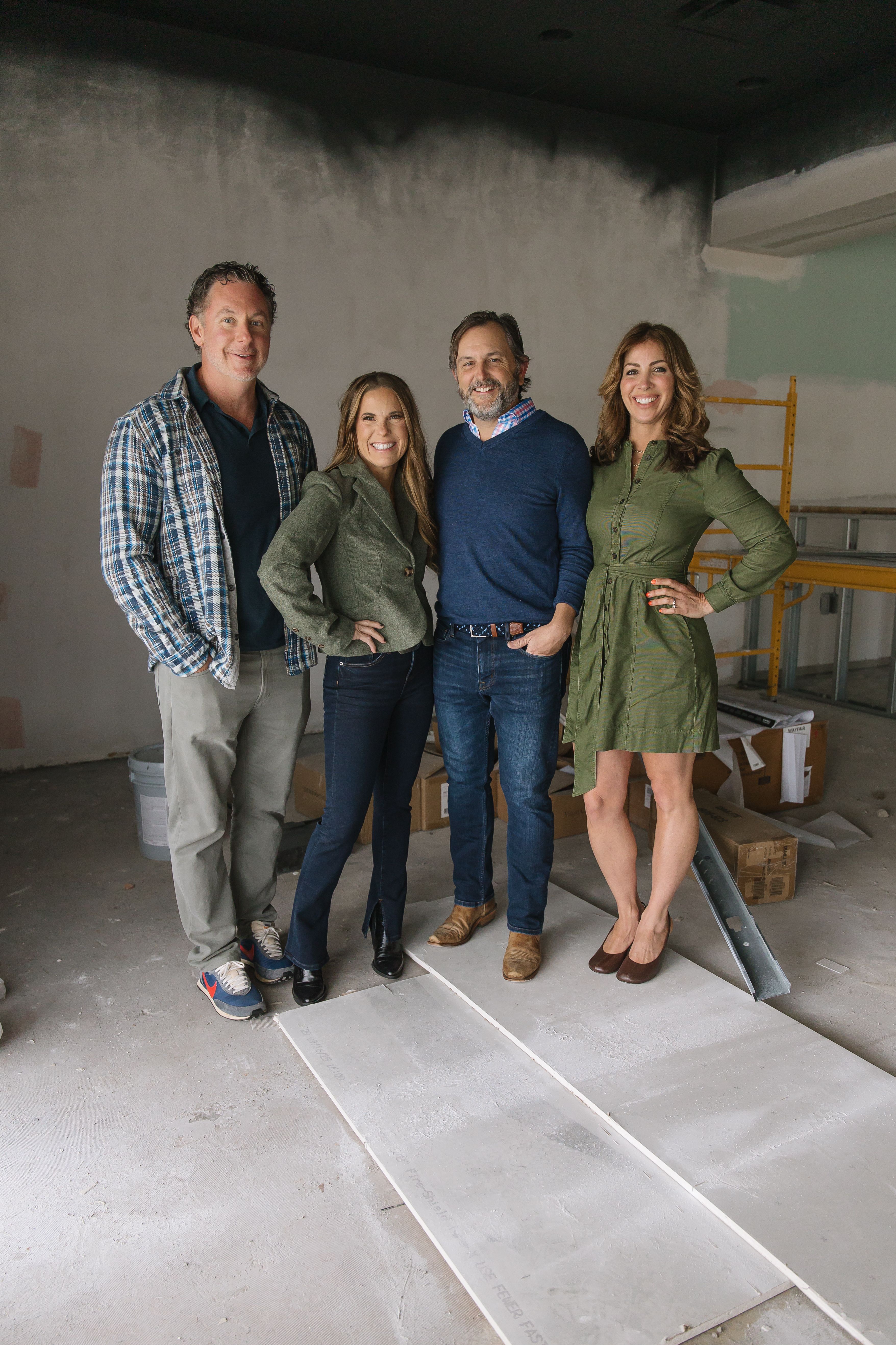 Four smiling adults standing in a partially finished room with construction materials; two men in casual shirts and jeans, two women in green outfits, one with a blazer, the other a dress.