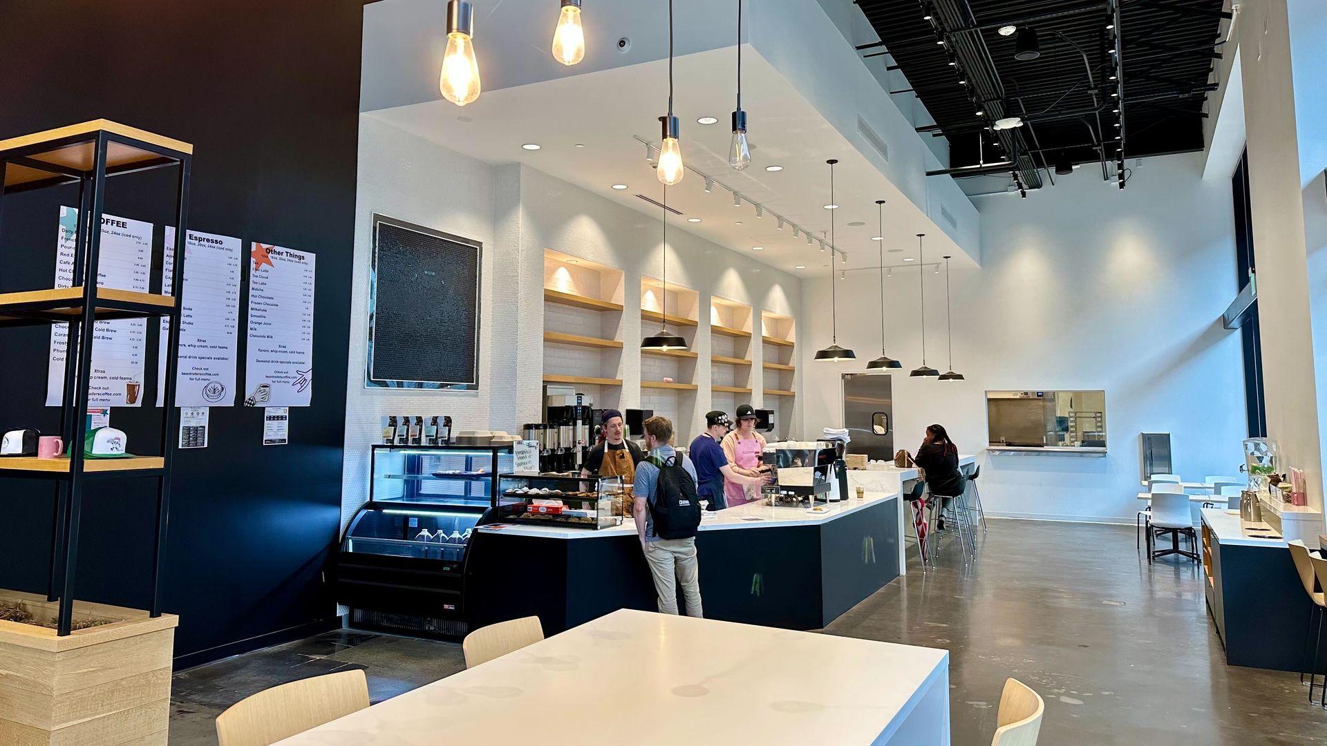 Bright modern cafe interior with white walls, black ceiling, and warm Edison bulbs. A long white counter with staff; navy wall with a chalkboard, glass display case, and light wood tables and chairs.