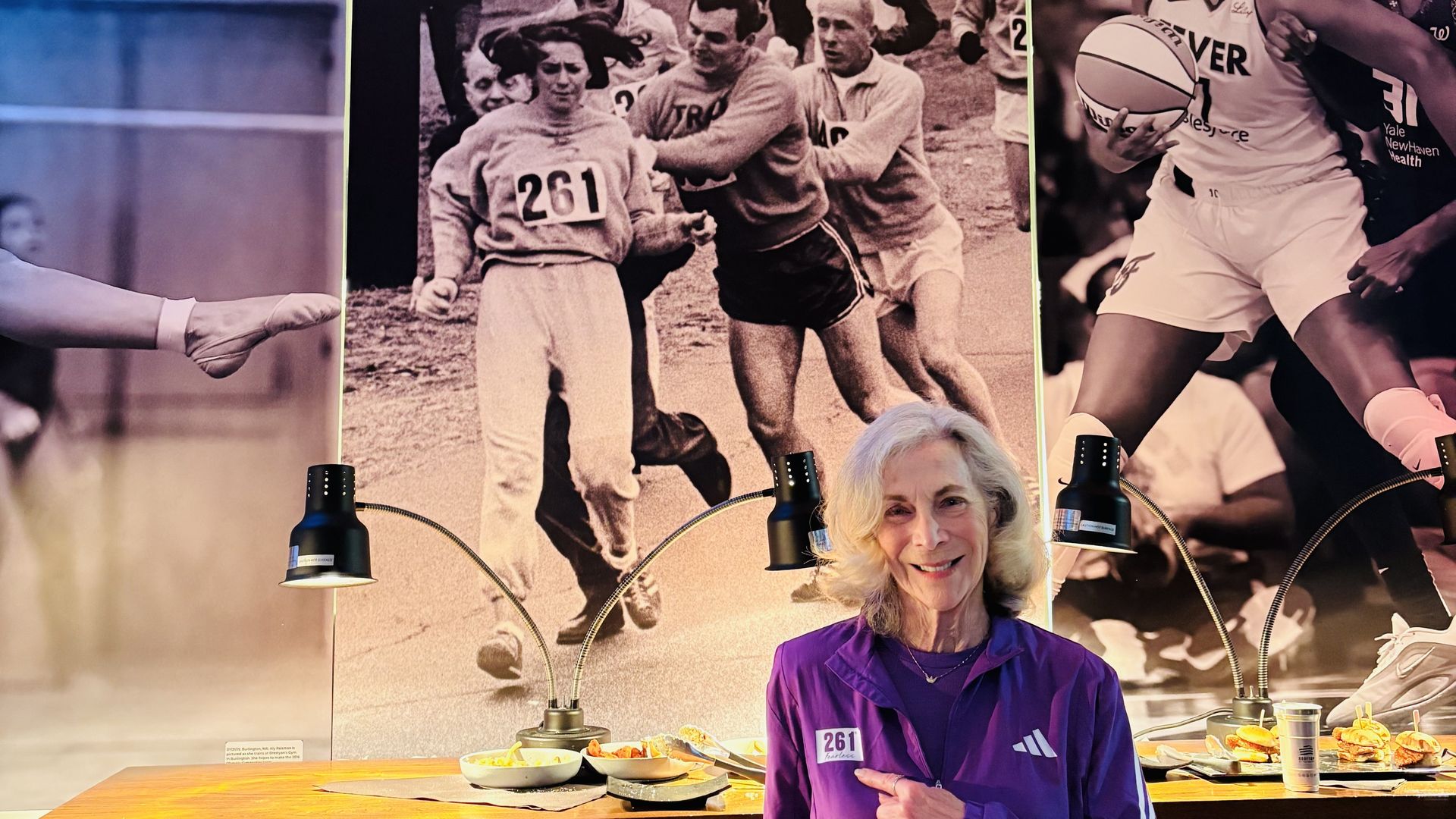 Katherine Switzer stands inside 211, a women's sports bar pop-up in Para Maria in Boston named after her historic 1967 Boston Marathon bib number. She points to her jacket, which has 261, as she stands in front of a large black-and-white photo of people trying to pull her out of the marathon route.