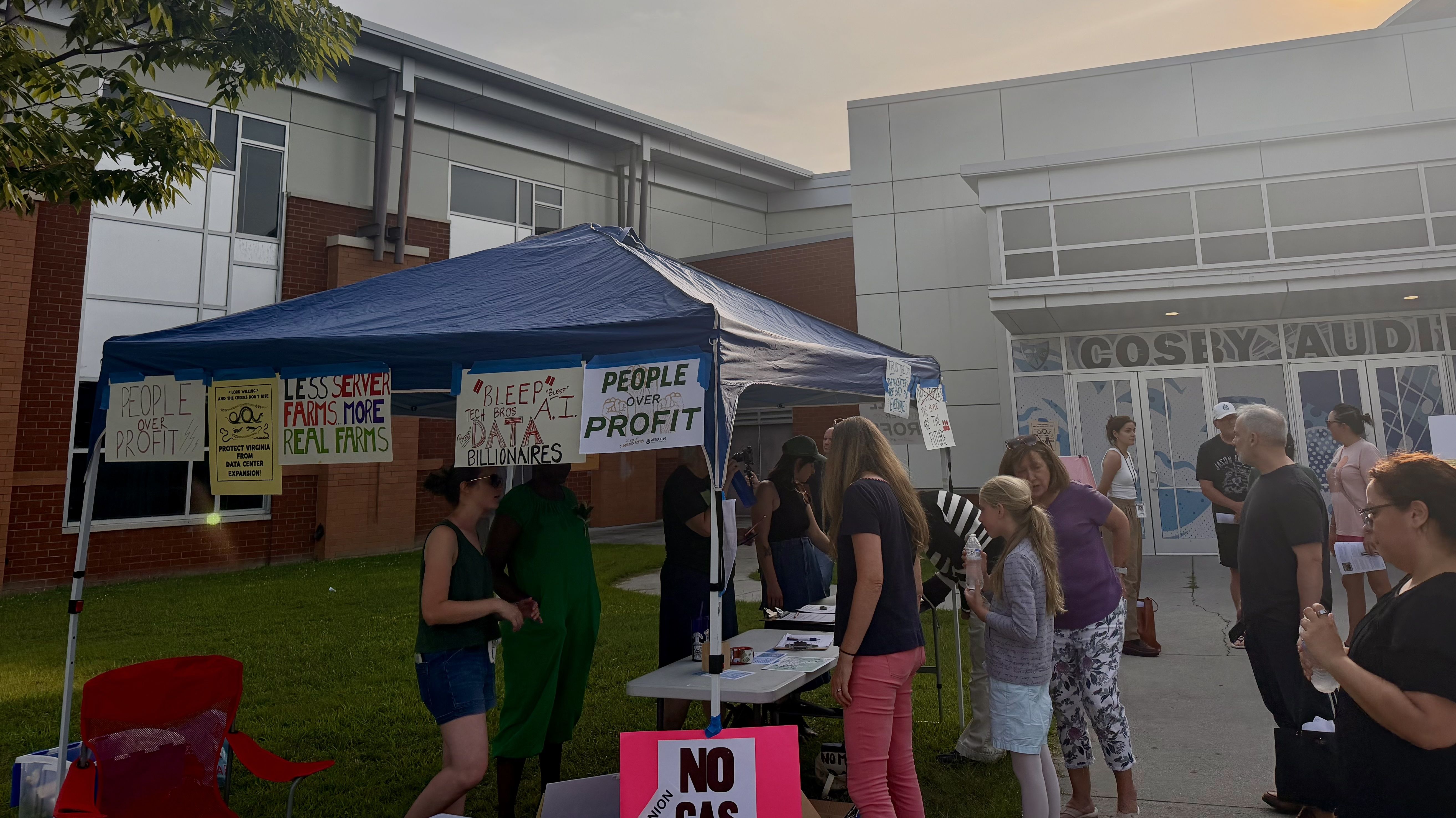 Blue canopy tent set up in a parking lot outside a modern brick-and-glass building. Posters read "PEOPLE OVER PROFIT" and "LESS SERVER FARMS, MORE REAL FARMS" as people wait in line.