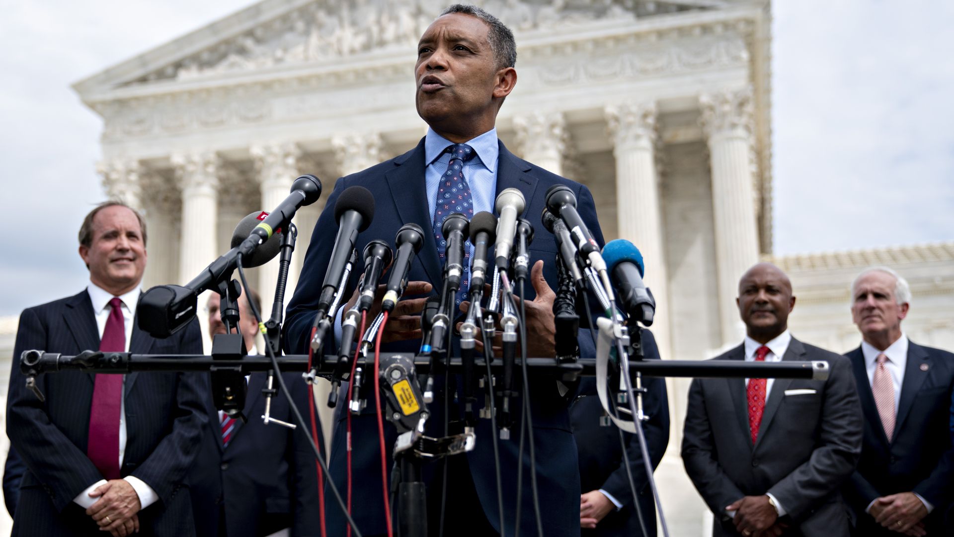 AG Racine speaking in a blue suit and tie with a dozen mics in front of him.