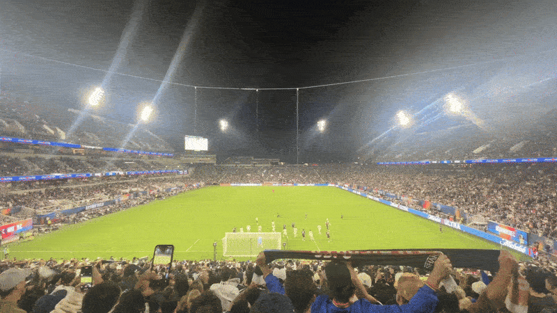 A view of a soccer stadium from the crowd behind the goal as a player scores. Blue smoke and fireworks go off as fans cheer. 