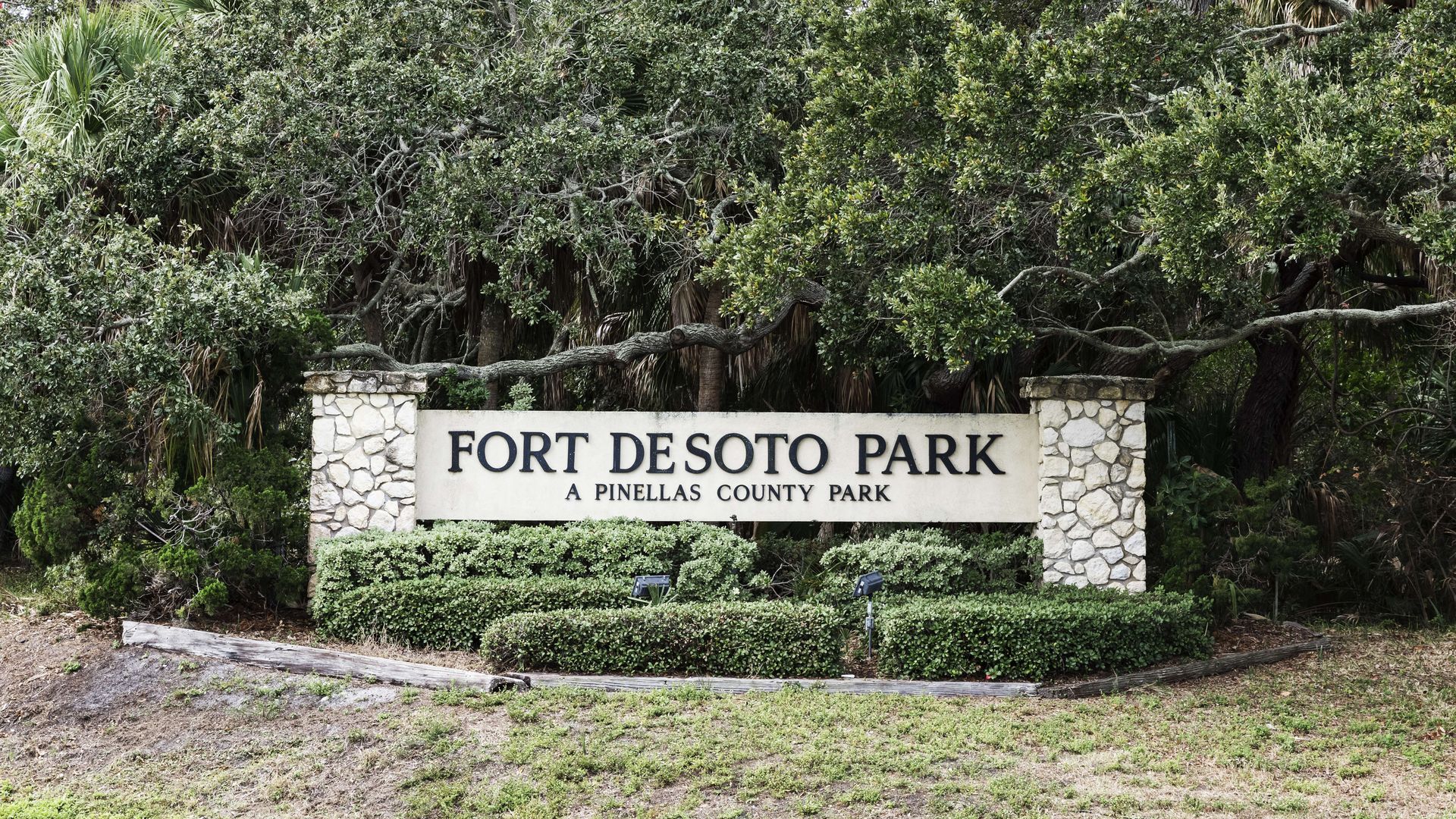 Fort De Soto Park sign with stone pillars, surrounded by dense trees and shrubs; a concrete panel reads "FORT DESOTO PARK" with "A PINELLAS COUNTY PARK" below.