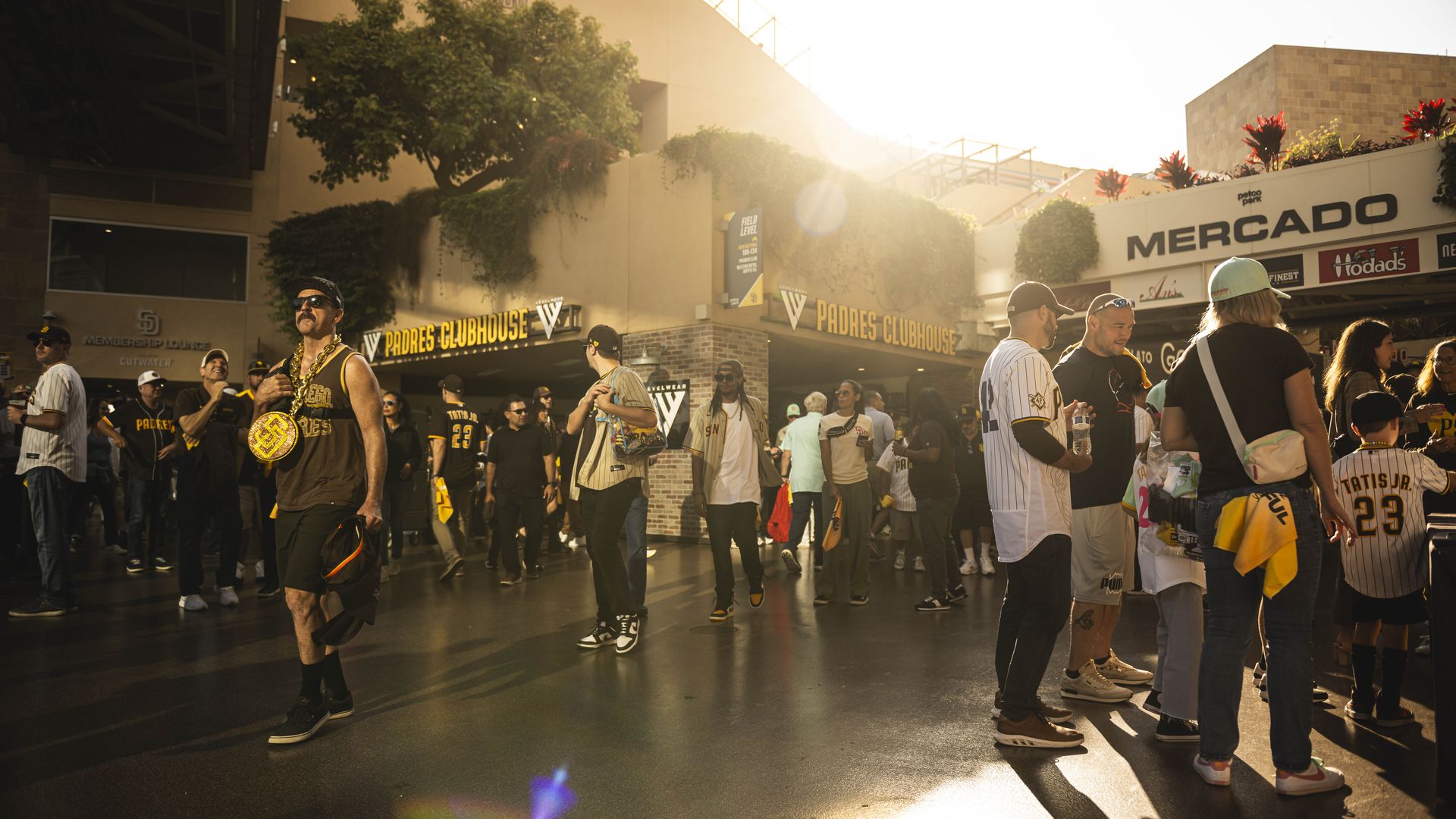 Fans dressed in San Diego Padres gear walk around the Petco Park concourse and stand in line at concessions vendors before a game as the sun sets. 