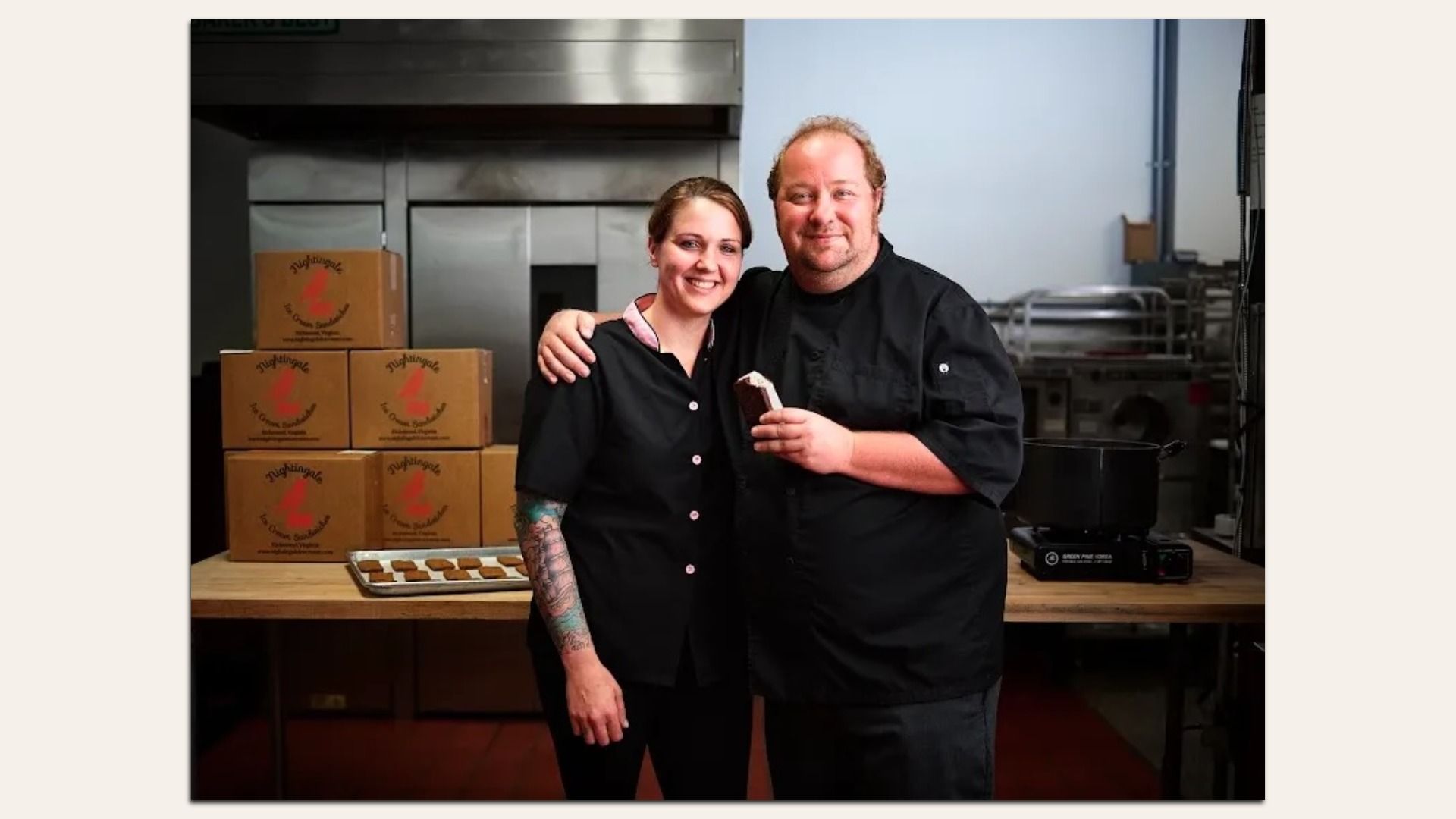 Two chefs, a tattooed-arm woman and a man, stand in a kitchen in black uniforms. The man holds a piece of cake; they smile. Behind them are boxes, a cookie tray, and stainless appliances.