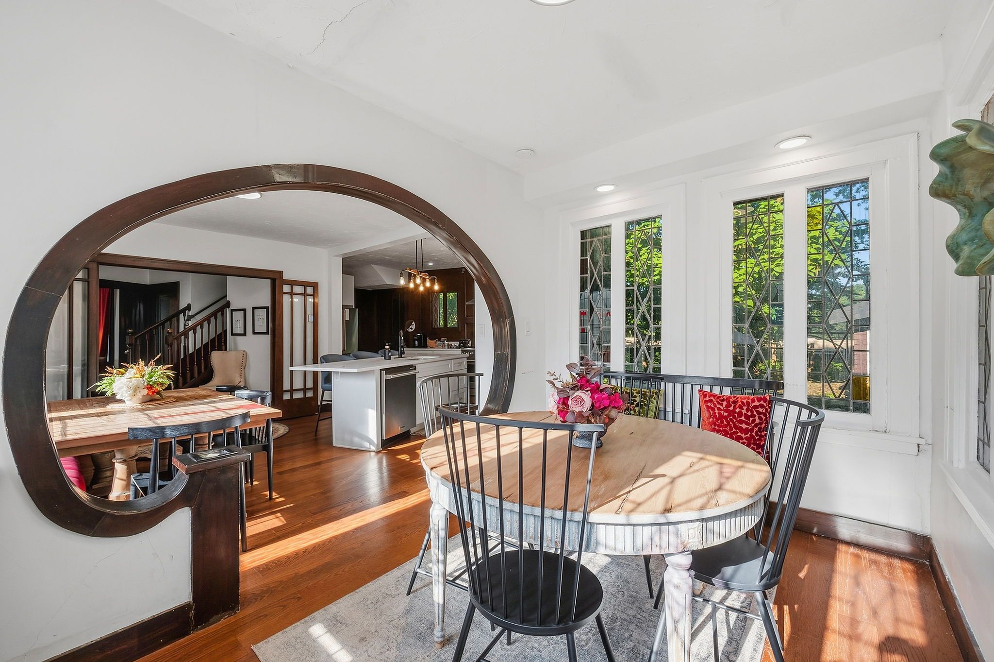 Bright dining area with a large circular dark wood frame on the left. Open kitchen in back, round wooden table with black metal chairs and a floral centerpiece. White walls, grid windows, greenery outside.