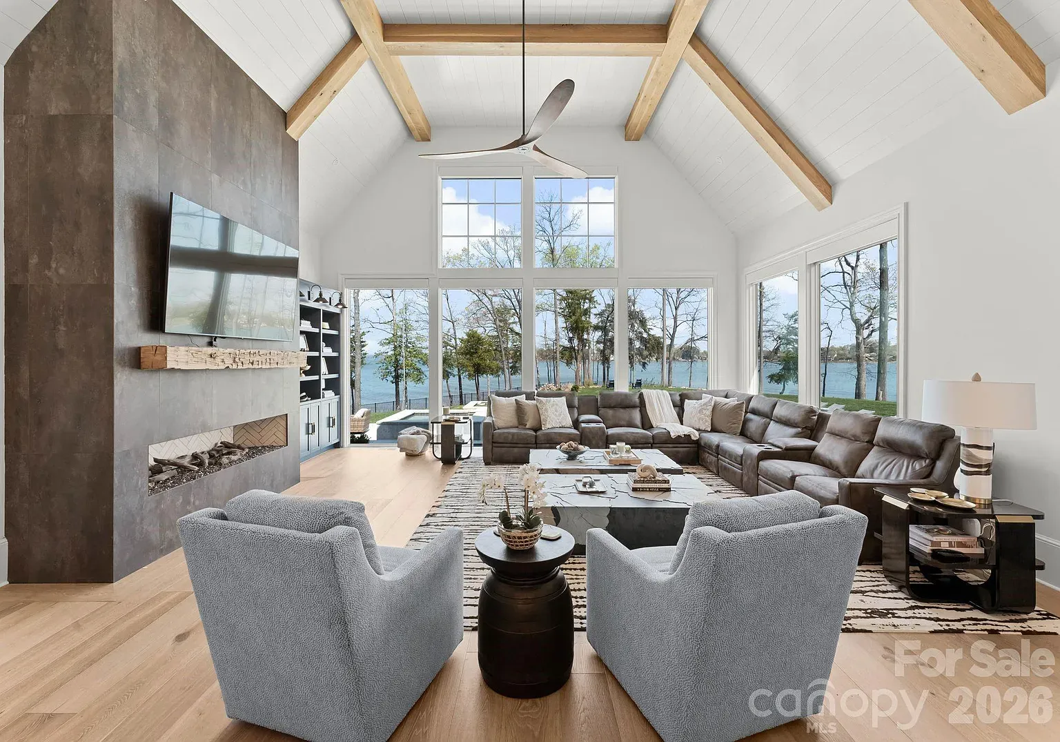 Bright living room with vaulted white ceiling and wooden beams, gray sectional sofa, two light-blue chairs, a stone fireplace with TV, and large windows framing a lake view.