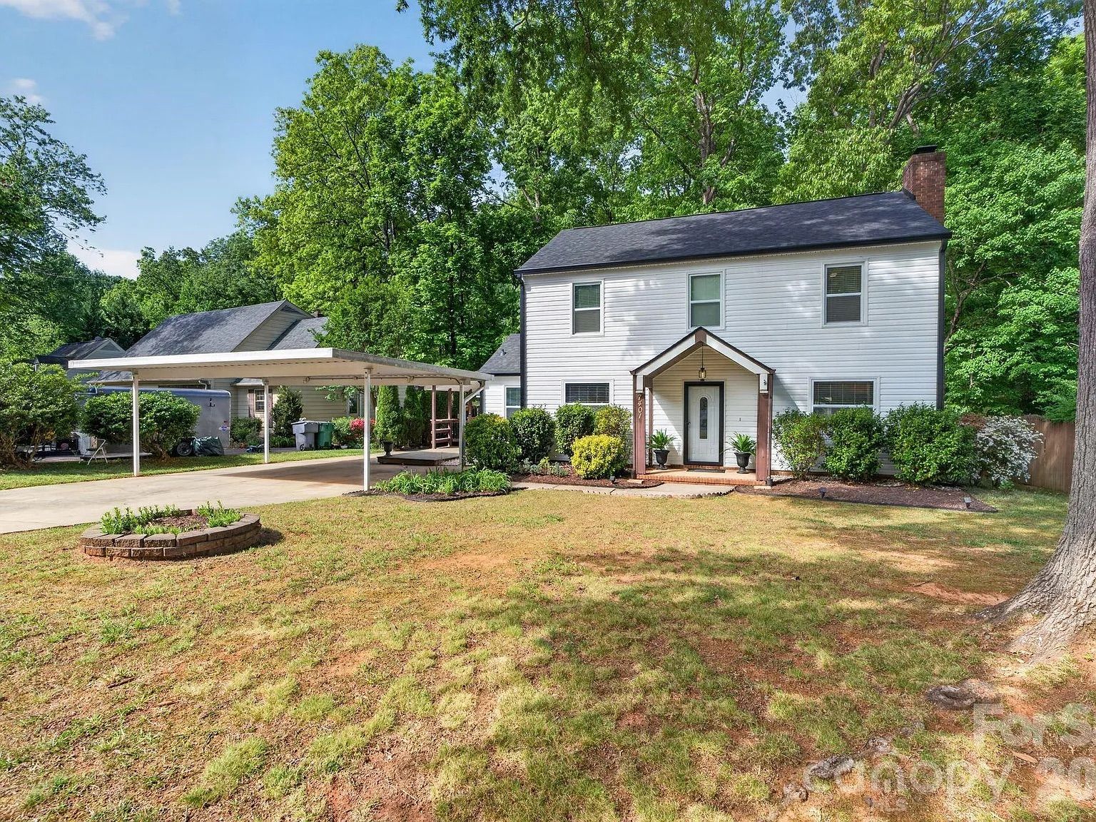 Two-story white house with dark roof, small front porch, and glass-panel door. Attached carport on left, paved driveway, circular brick flower bed, and green shrubs; tall trees and blue sky behind.