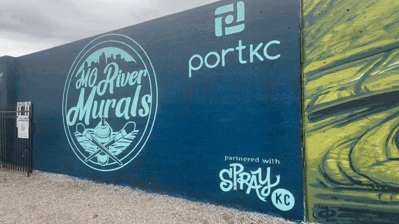Blue mural wall featuring circular logo "Mo River Murals" with paintbrush and spray can, alongside text "portkc" and "partnered with Spray KC" in light blue. Adjacent yellow graffiti visible.