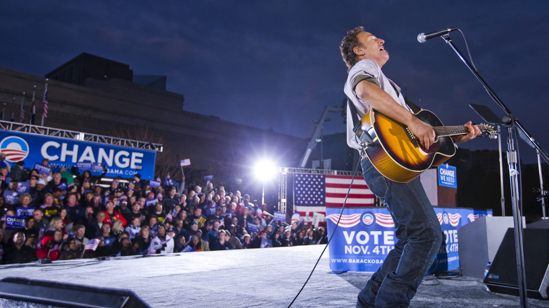 Male singer with a sunburst acoustic guitar performs on an outdoor stage at dusk; a large crowd holds Change We Need and Vote signs with American flags in the background.
