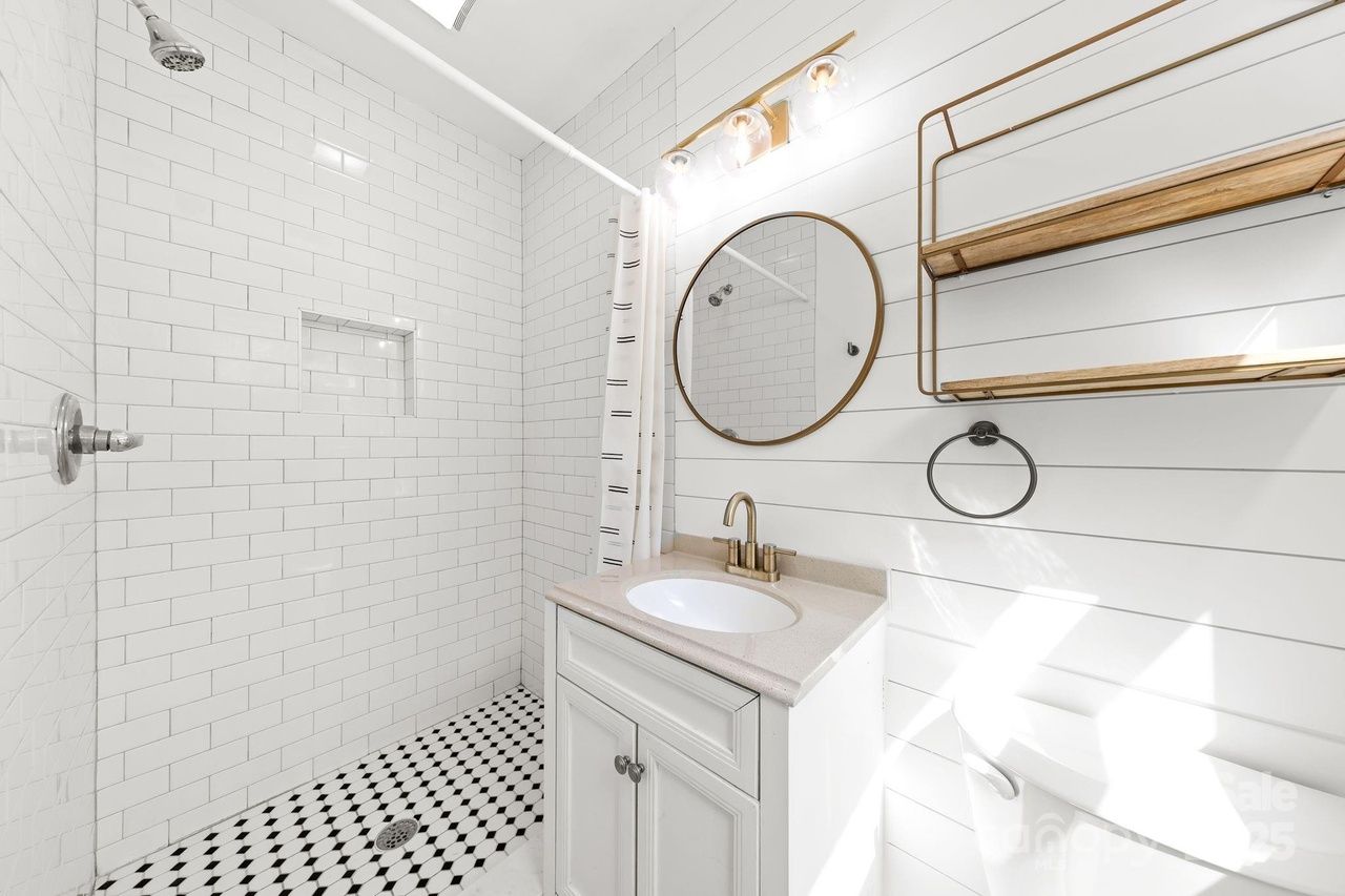 Bright white bathroom with subway tile shower, black and white patterned floor, round mirror with bronze frame, white vanity with beige countertop, and bronze fixtures.