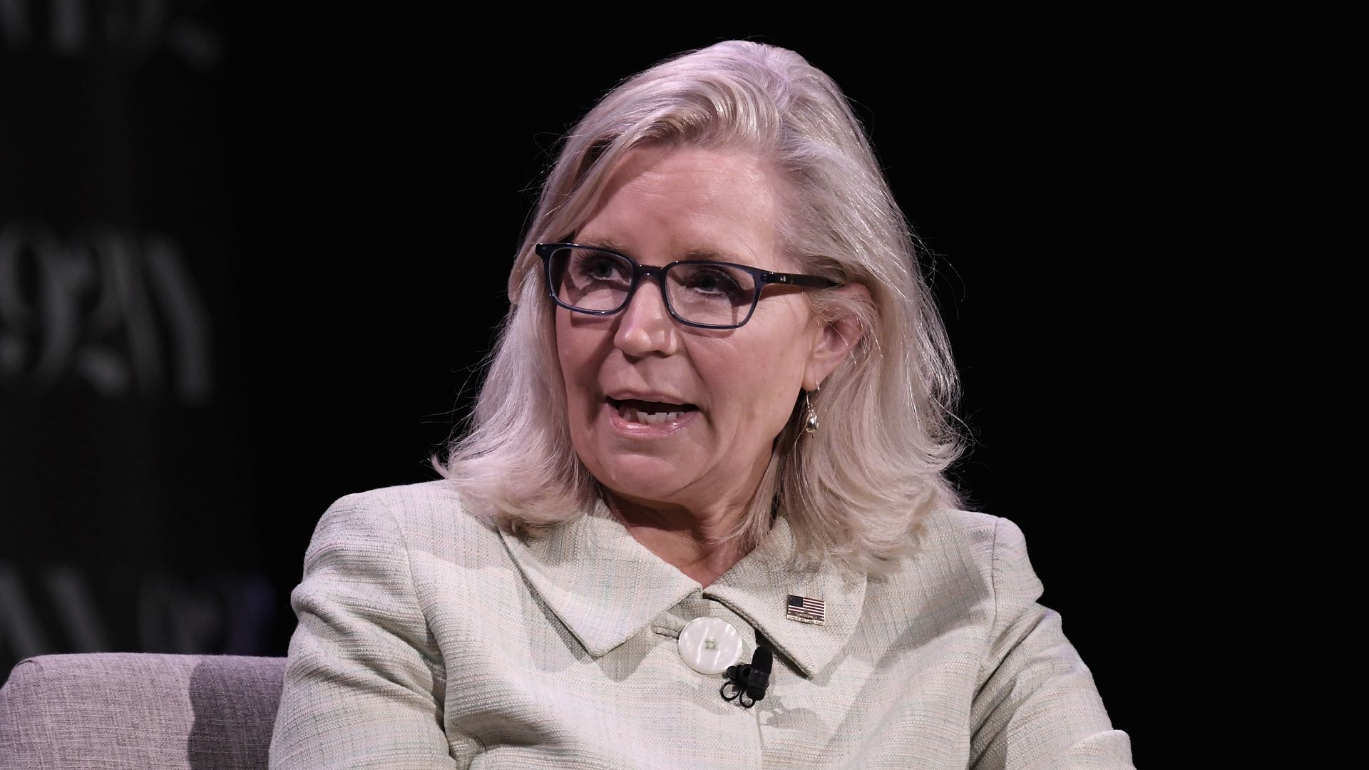 Liz Cheney speaks with an American flag pinned on her lapel and a dark background. She is seated. 