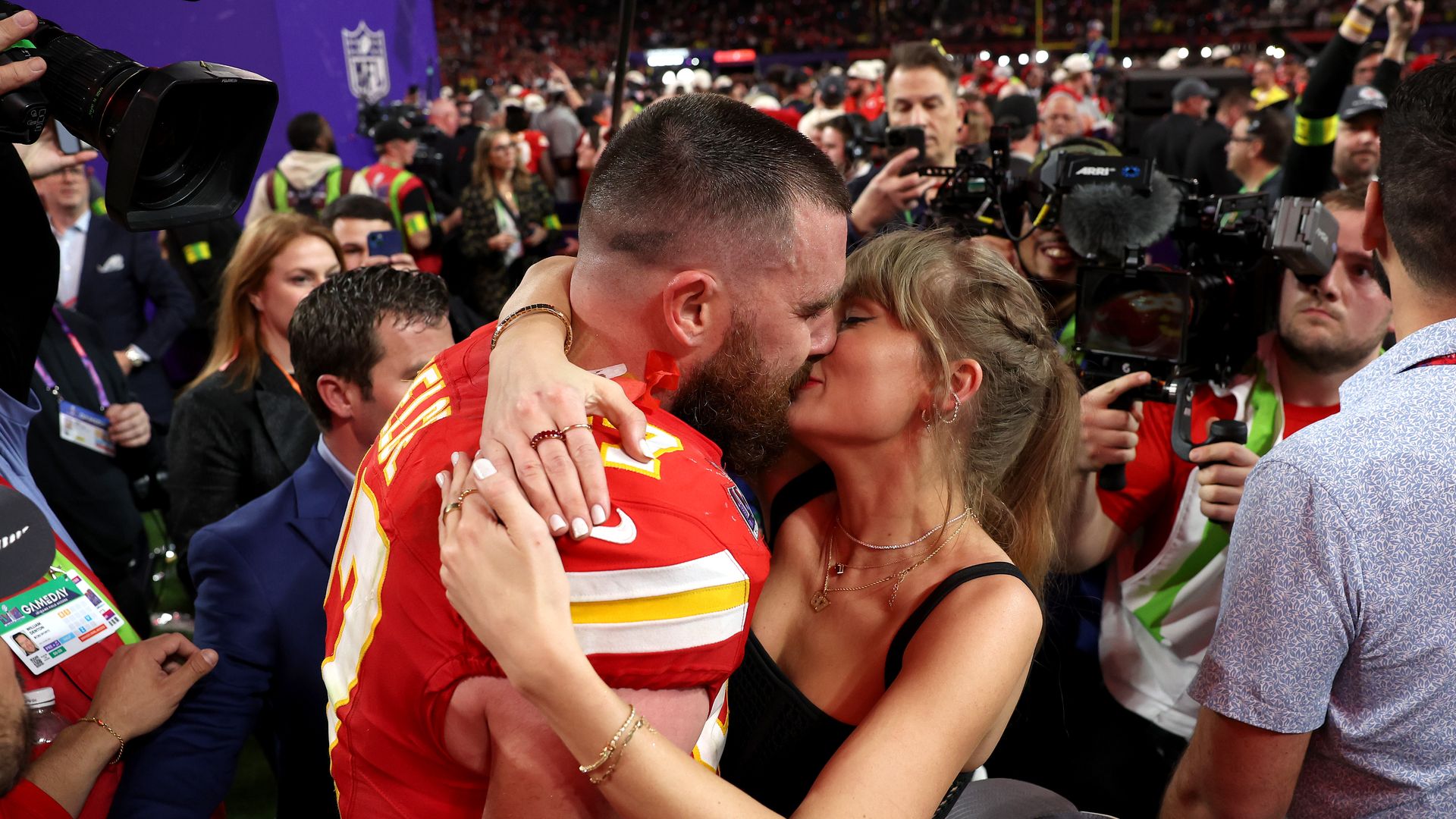 Football player Travis Kelce in red uniform kissing Taylor Swift in a black dress amid a crowd with cameras and media in the background at a sports event.
