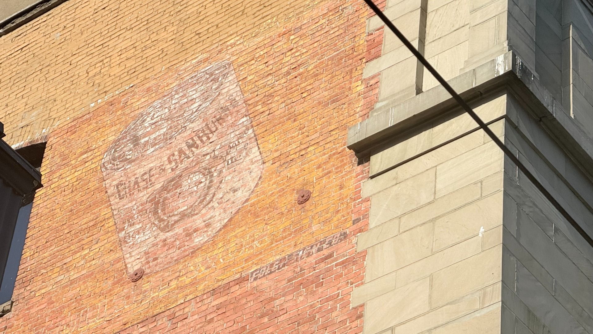 Orange brick wall with a large faded circular mural or logo. Beige stone building corner on the right; dark diagonal wires cross the scene.