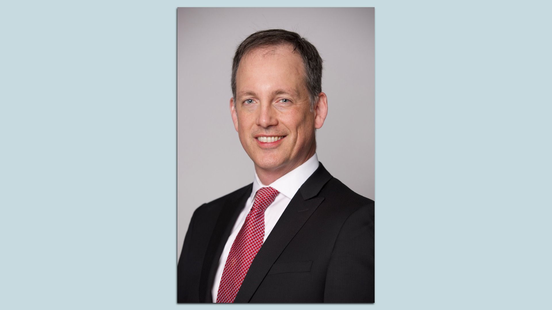 An individual wearing a suit with a red tie smiles and poses for a headshot