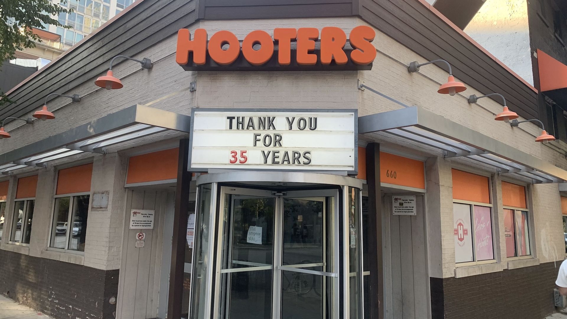 Exterior corner of a Hooters restaurant with bright orange HOOTERS letters on a beige wall, orange wall lamps, and a marquee that reads "THANK YOU FOR 35 YEARS".
