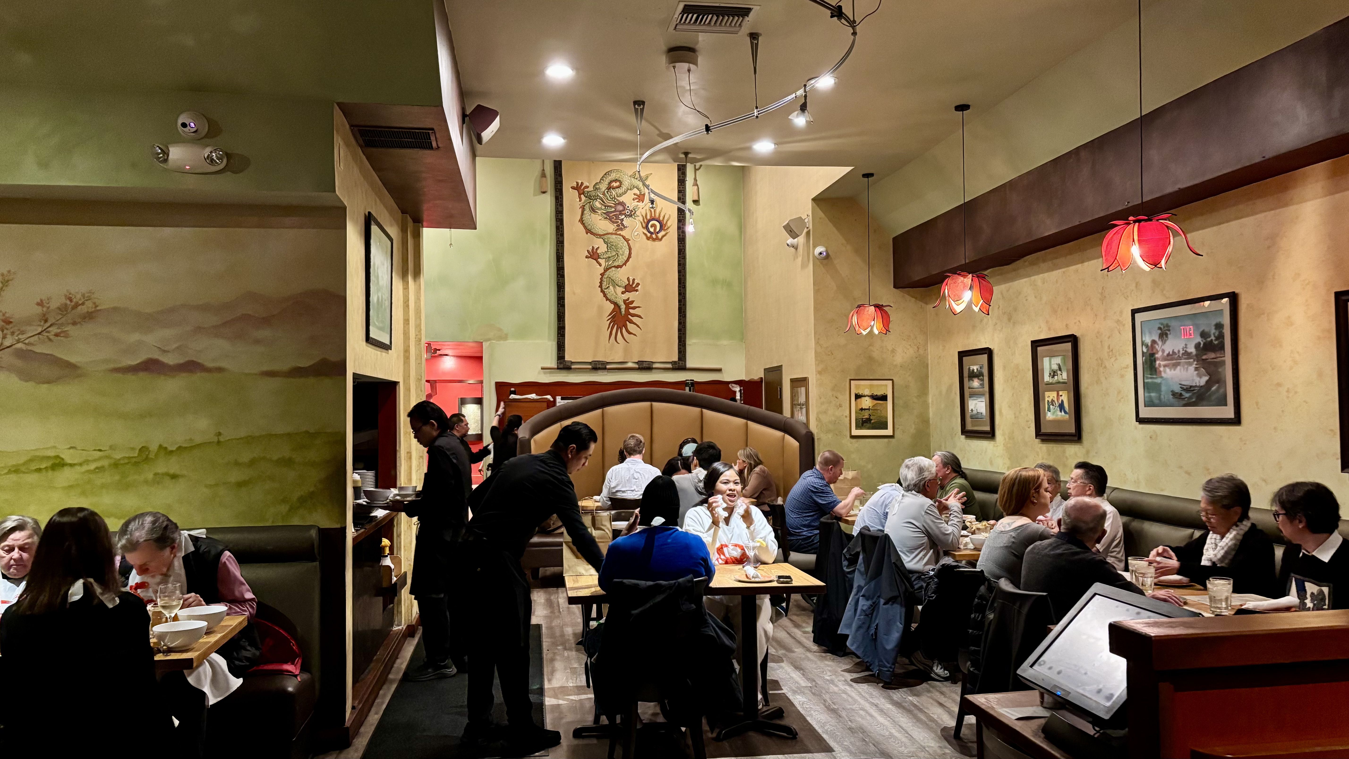 Interior of a warmly lit restaurant with several diners seated at tables and booths, red flower-shaped hanging lamps, a dragon mural on the back wall, and framed pictures on the side walls.