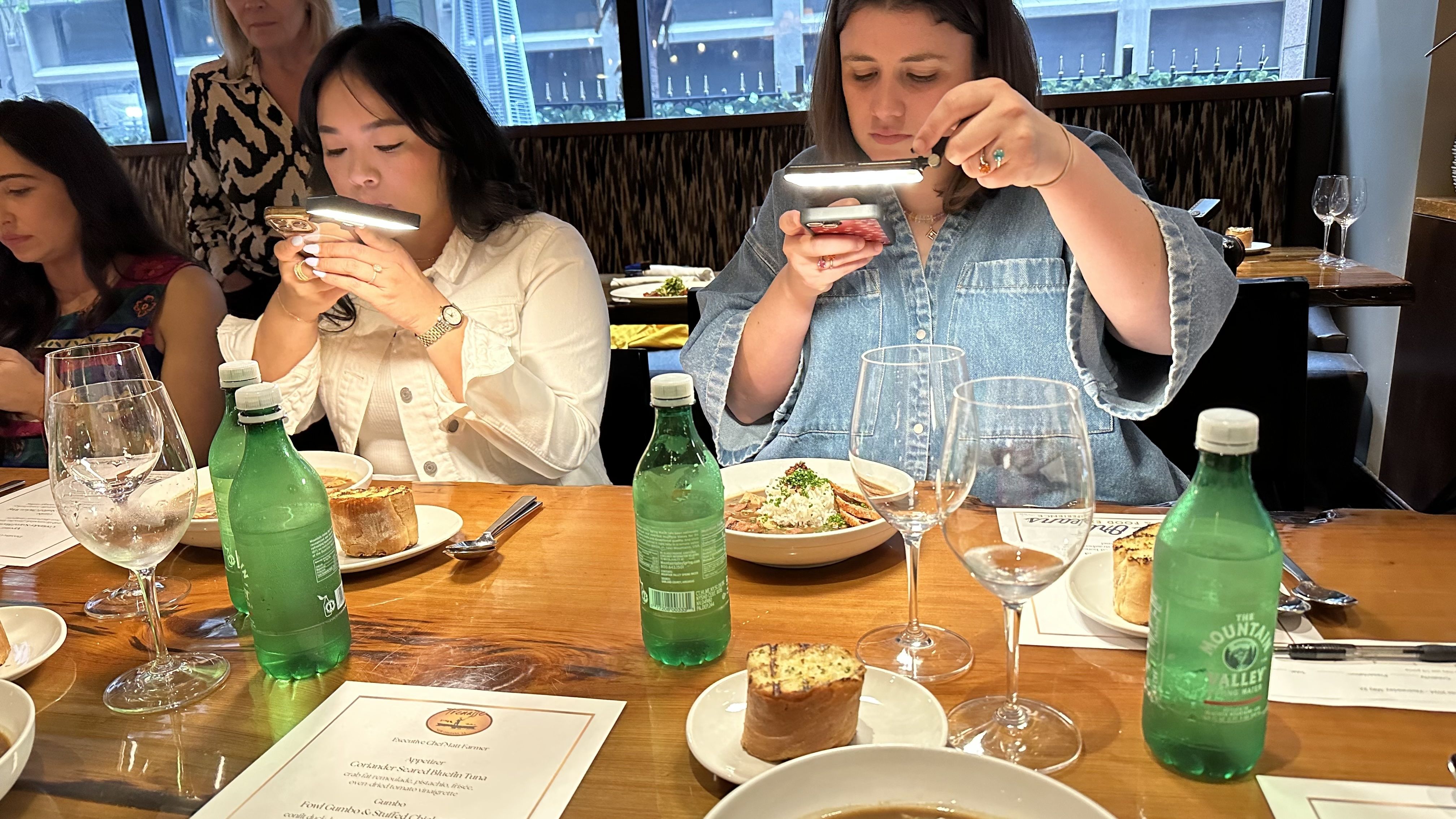Two women light their plates as they sit at a table and take photos of their meals.