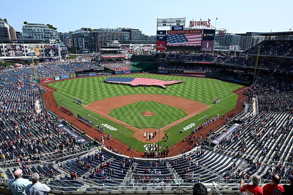 A photo shows a birds-eye-view of a baseball stadium with a large American flag spreading out across the field, being held by people.