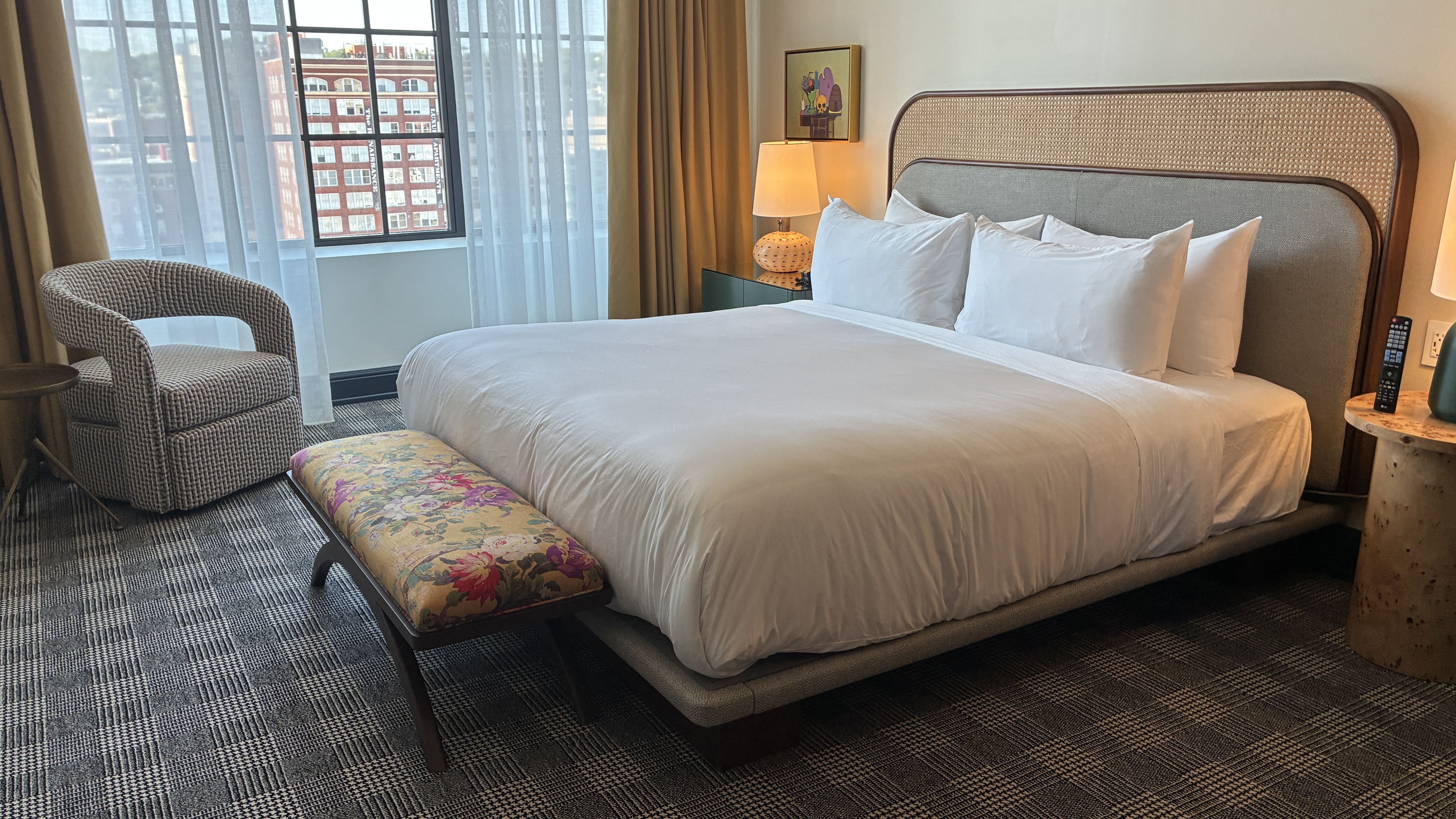 Hotel room with a large bed, curved wicker headboard, and white linens. Floral bench at the foot, checkered carpet, a chair by the window, and a nightstand with lamp and artwork.