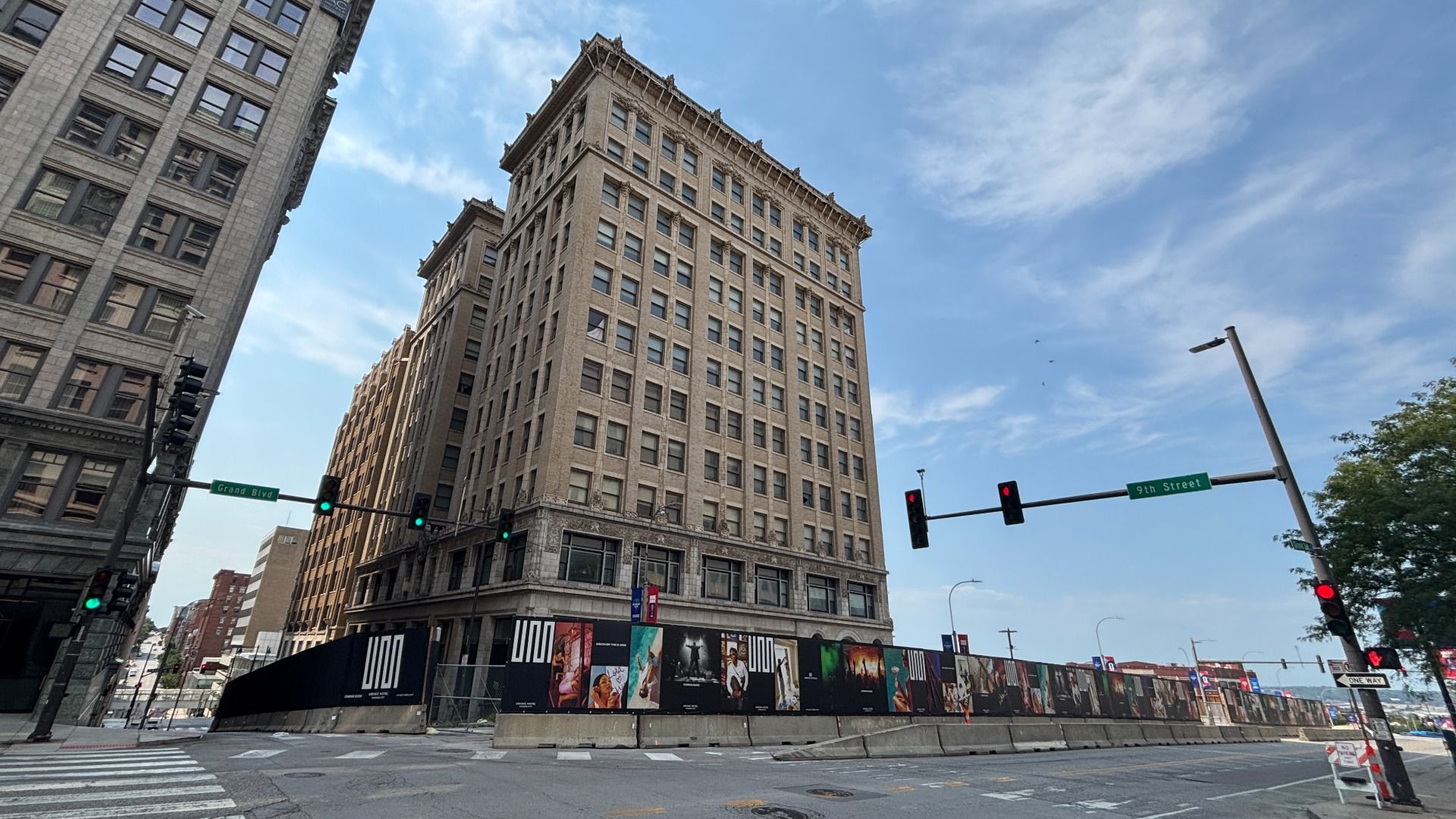 City street scene with tall beige brick buildings and ornate cornices against a bright blue sky. Corner shows Grand Blvd and 9th Street signs, traffic lights, crosswalk, and a low construction barrier with posters.