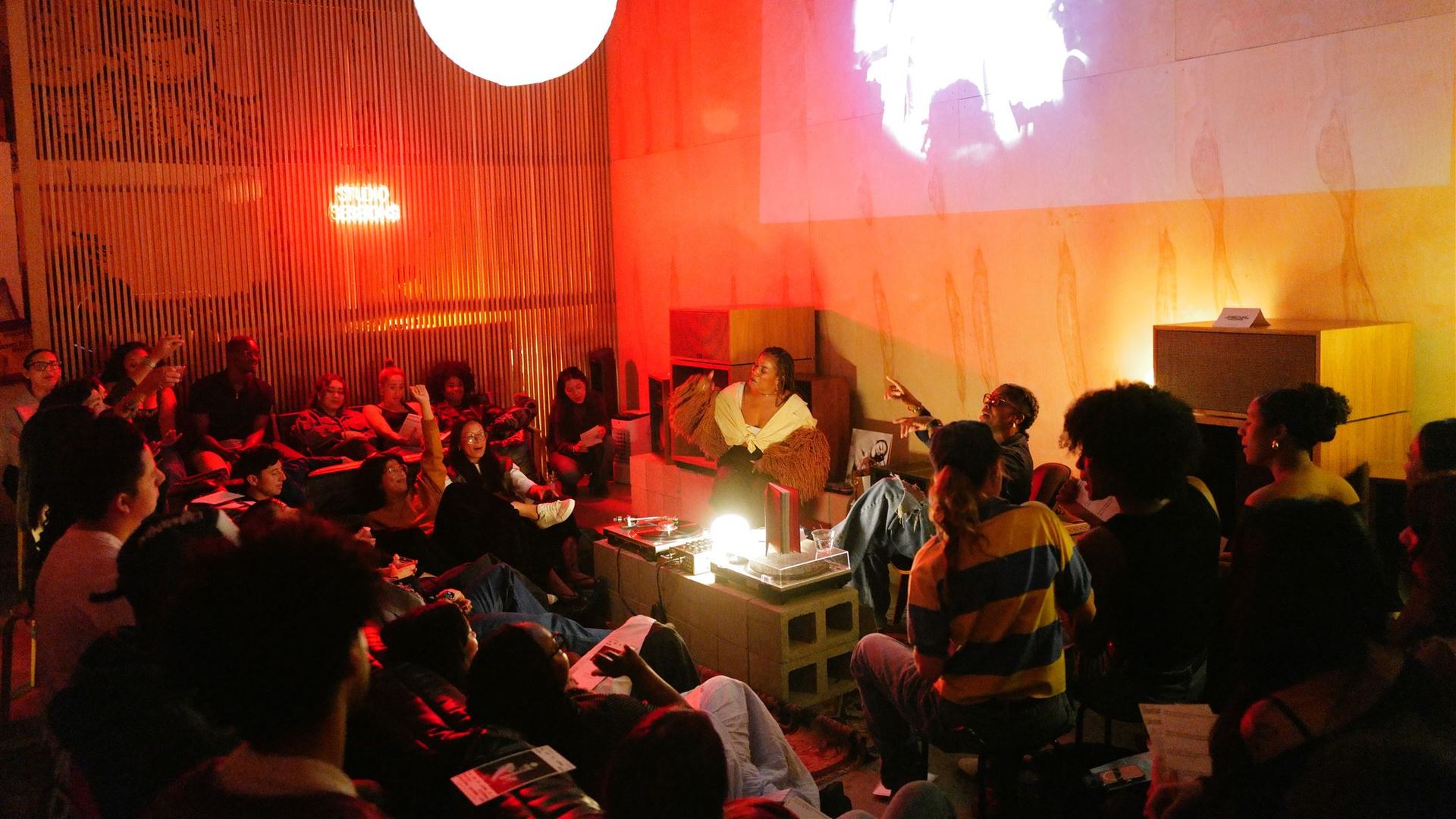 Crowded, red-lit room with a performer in fringe on a small stage; audience sits around, some raising hands. A large white ball light hangs left, and a black-and-white projection covers the wall.