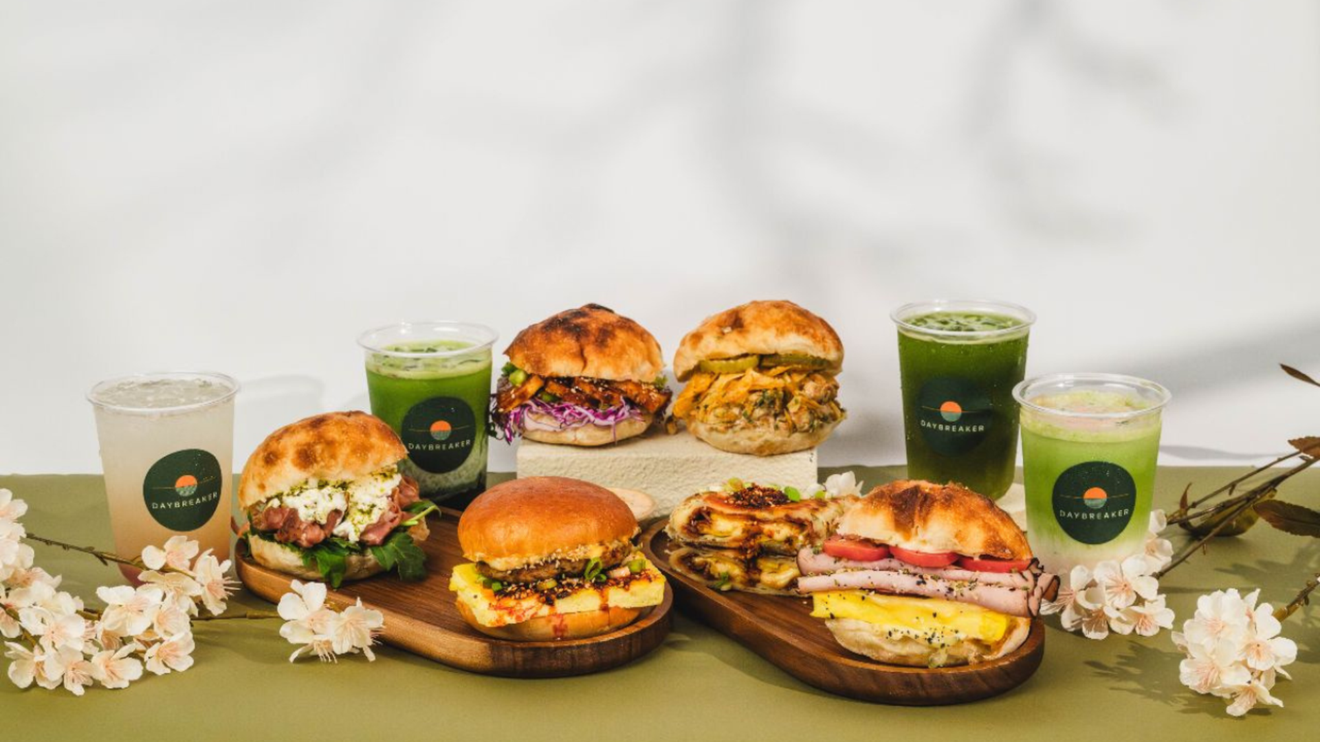 Assorted burgers and sandwiches on wooden boards with green Daybreaker cups and pale beverages, framed by pink blossoms on a light backdrop.