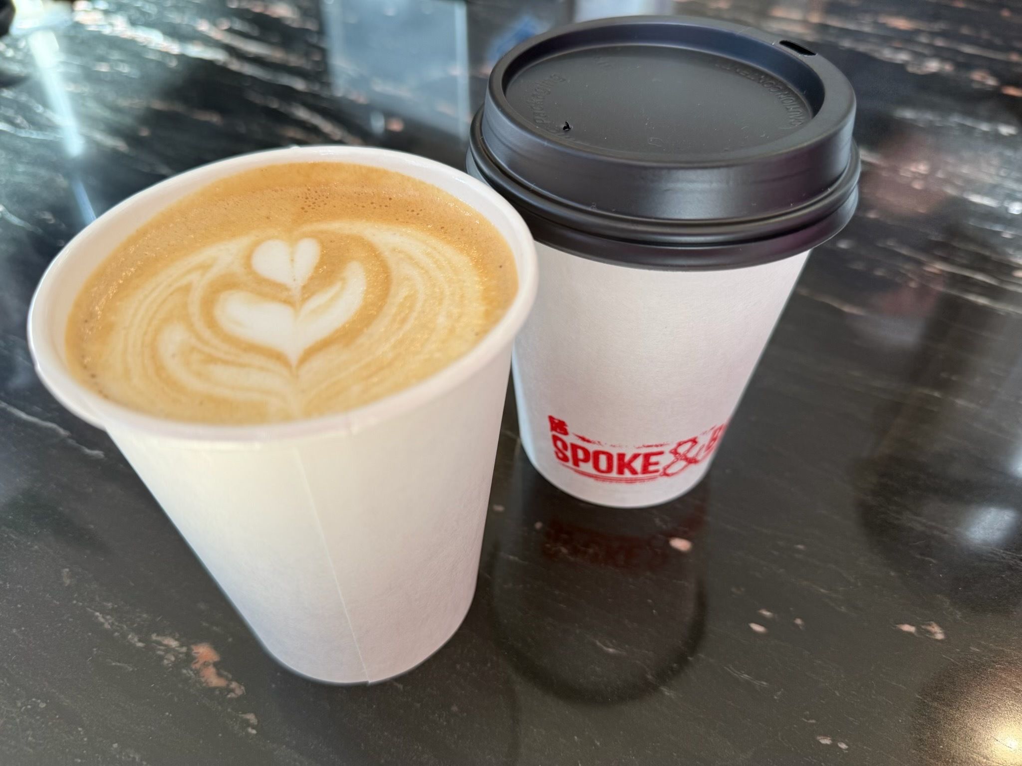 Two coffee cups on a dark marble table: a latte with heart-shaped foam art in a white cup on the left, and a take-away cup with a black lid and sleeve on the right.