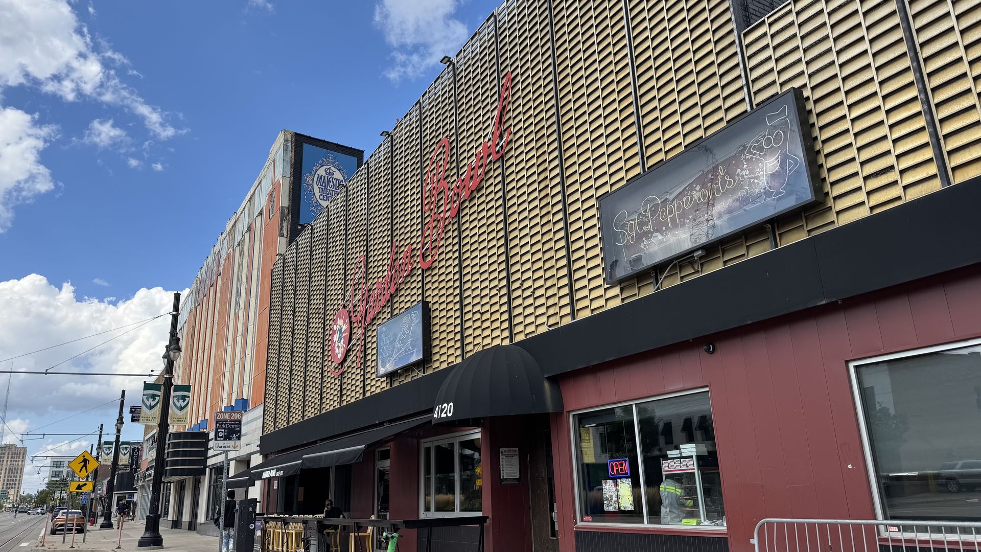 Street view of a red storefront with a gold lattice facade and a large red cursive sign. A blue circular sign reads "The Majestic Theatre Center." Another sign reads "Garden Bowl" in giant letters. Bright blue sky with a few clouds; outdoor seating under awnings.