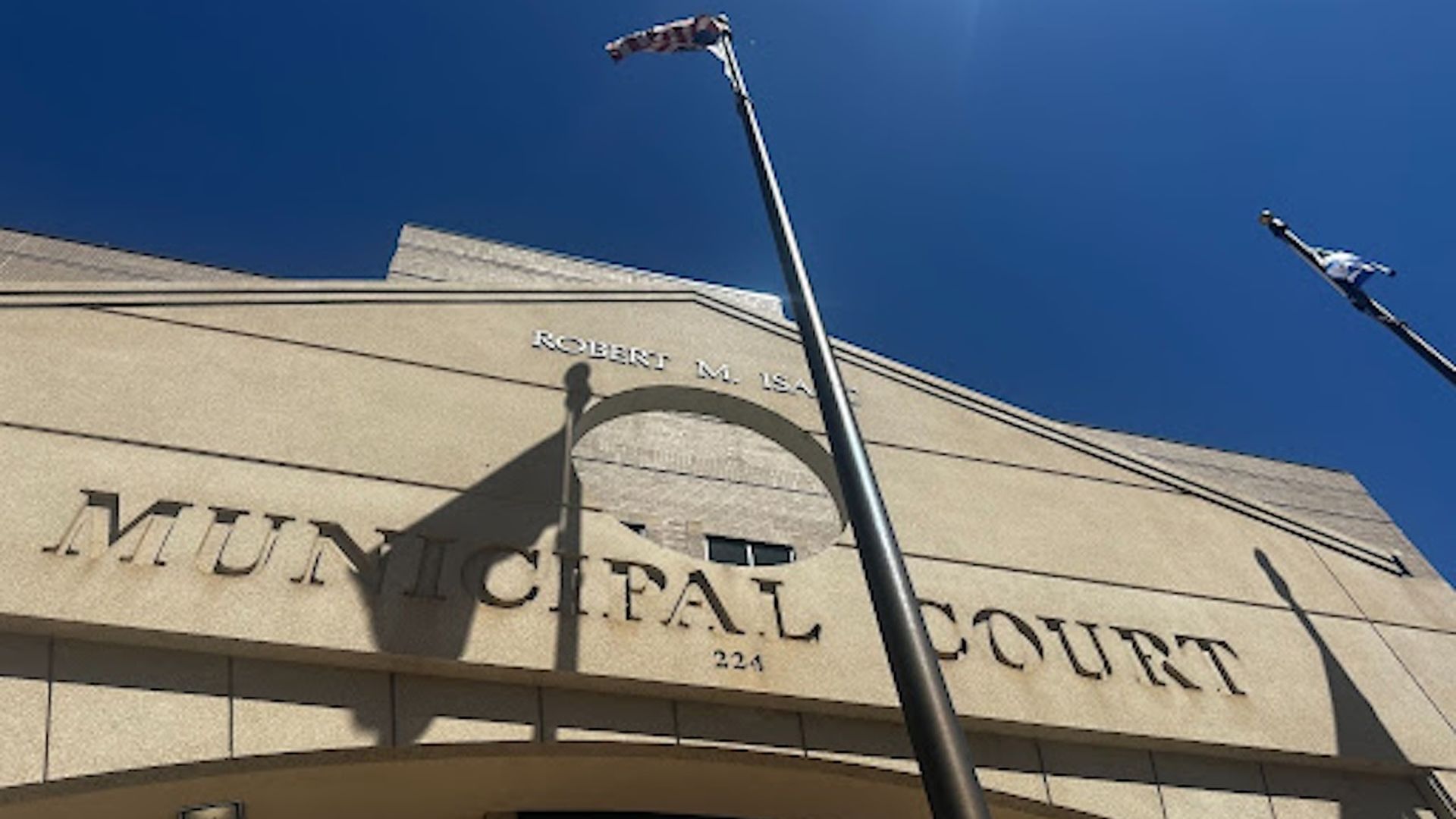 Beige municipal court building with large "MUNICIPAL COURT" lettering, a bright blue sky, and two flagpoles with fluttering flags; sunlit entrance and curved archway.
